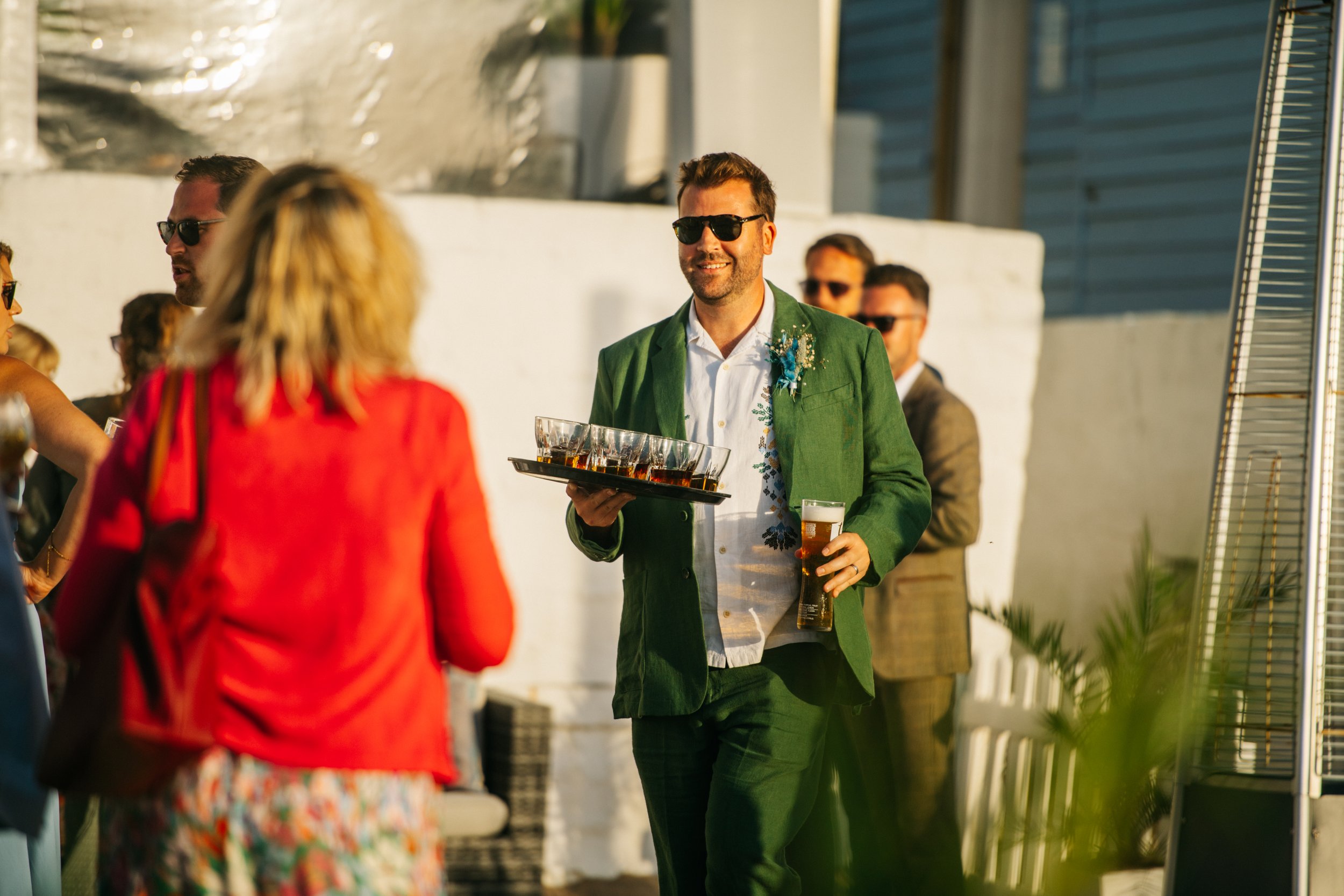 A man in a green suit holding a tray of glasses and a beer glass at an outdoor social gathering.