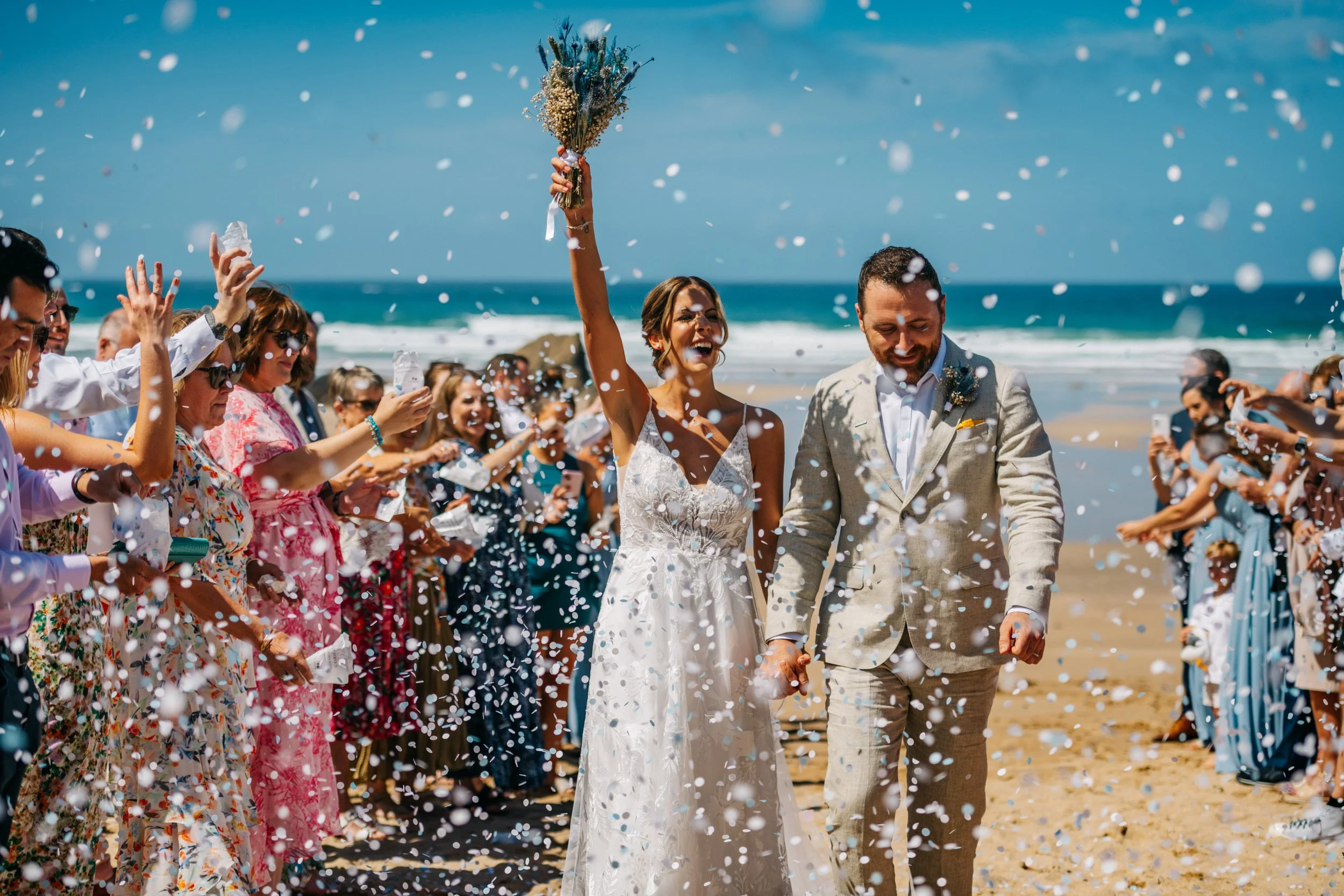 A newlywed couple walking hand in hand on the beach while guests throw confetti and celebrate.