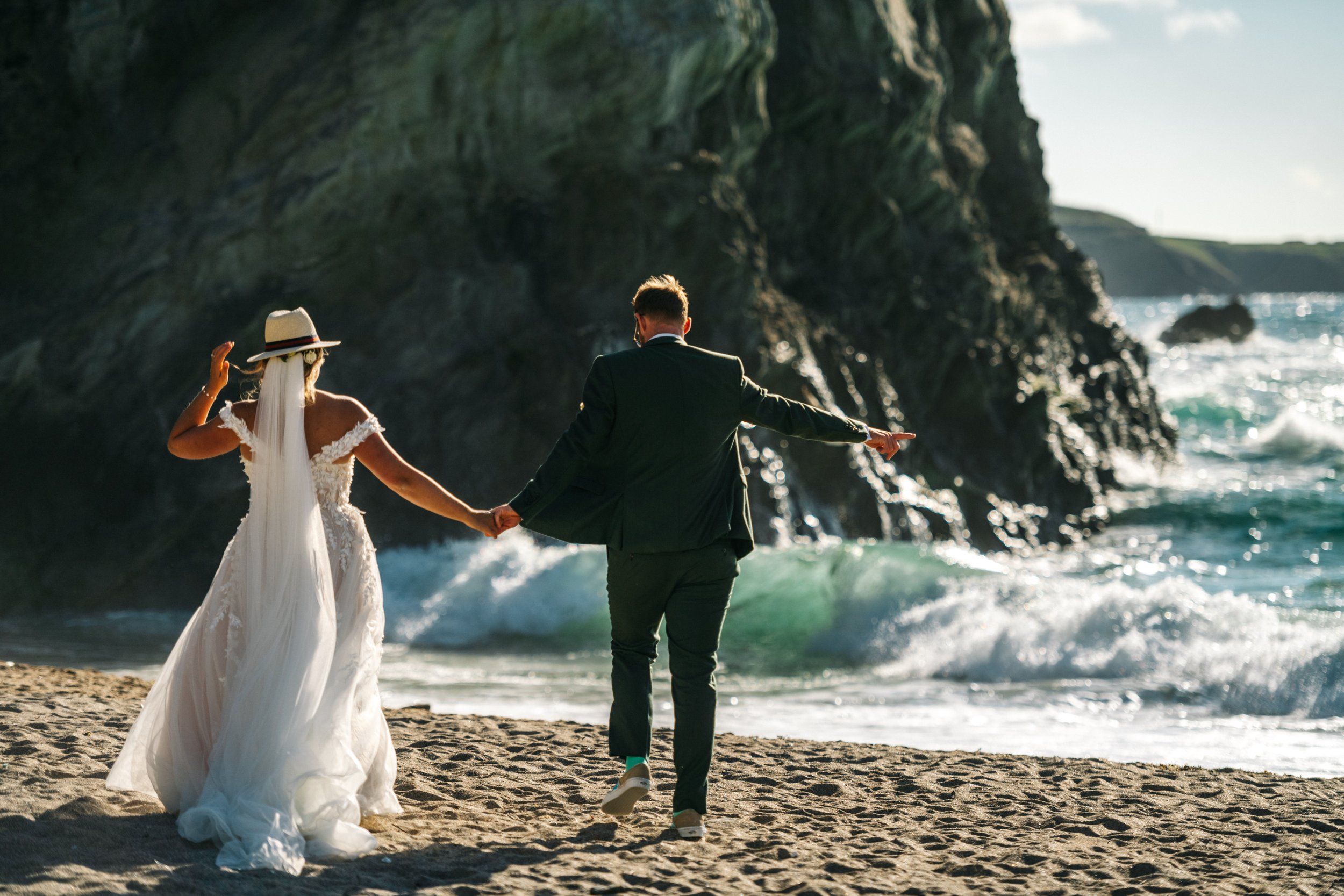 A bride and groom walking hand-in-hand on a beach with cliffs in the background, holding hands, during sunset or sunrise.