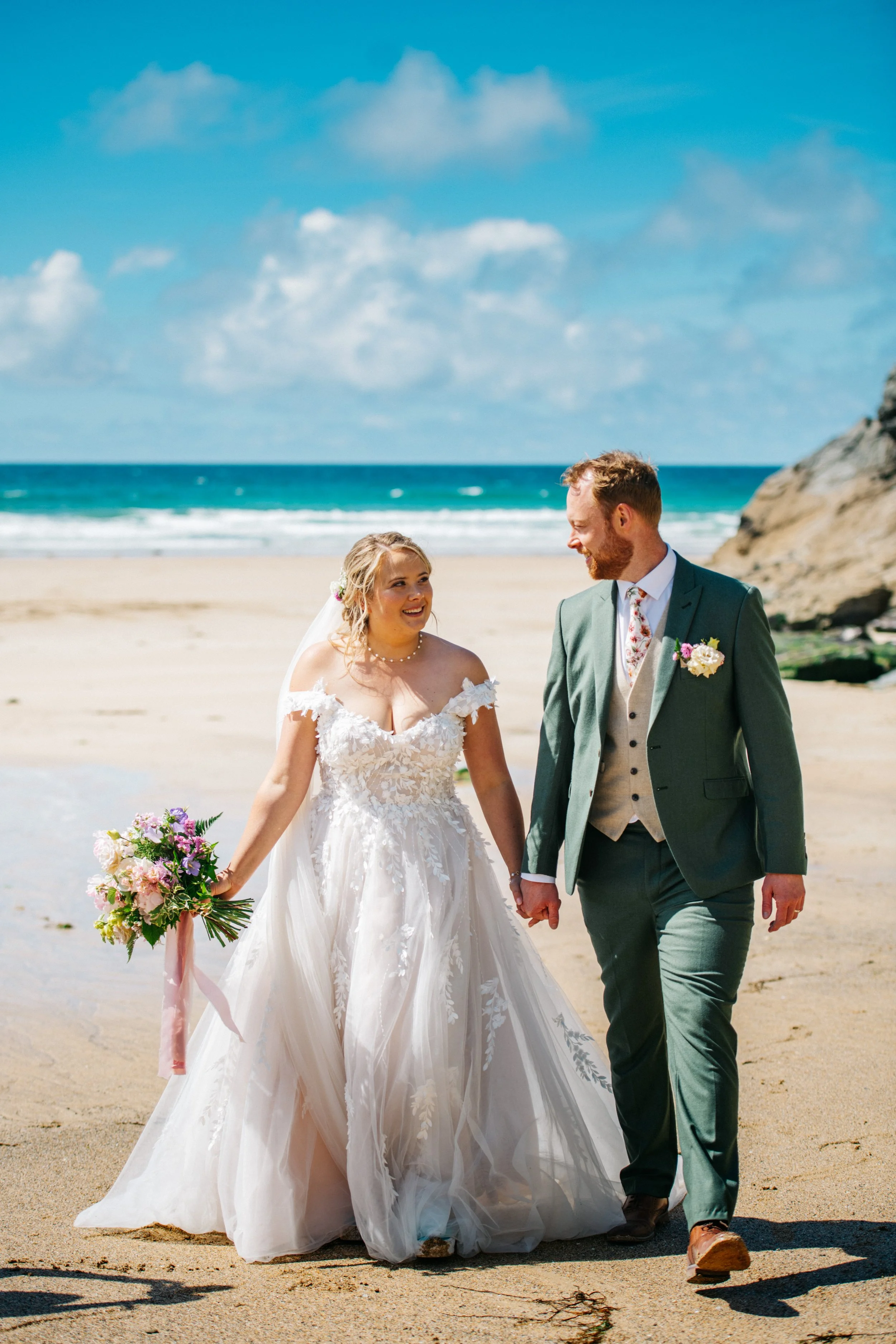 A bride and groom walking hand in hand on a beach, smiling at each other, with the ocean and a blue sky with clouds in the background.