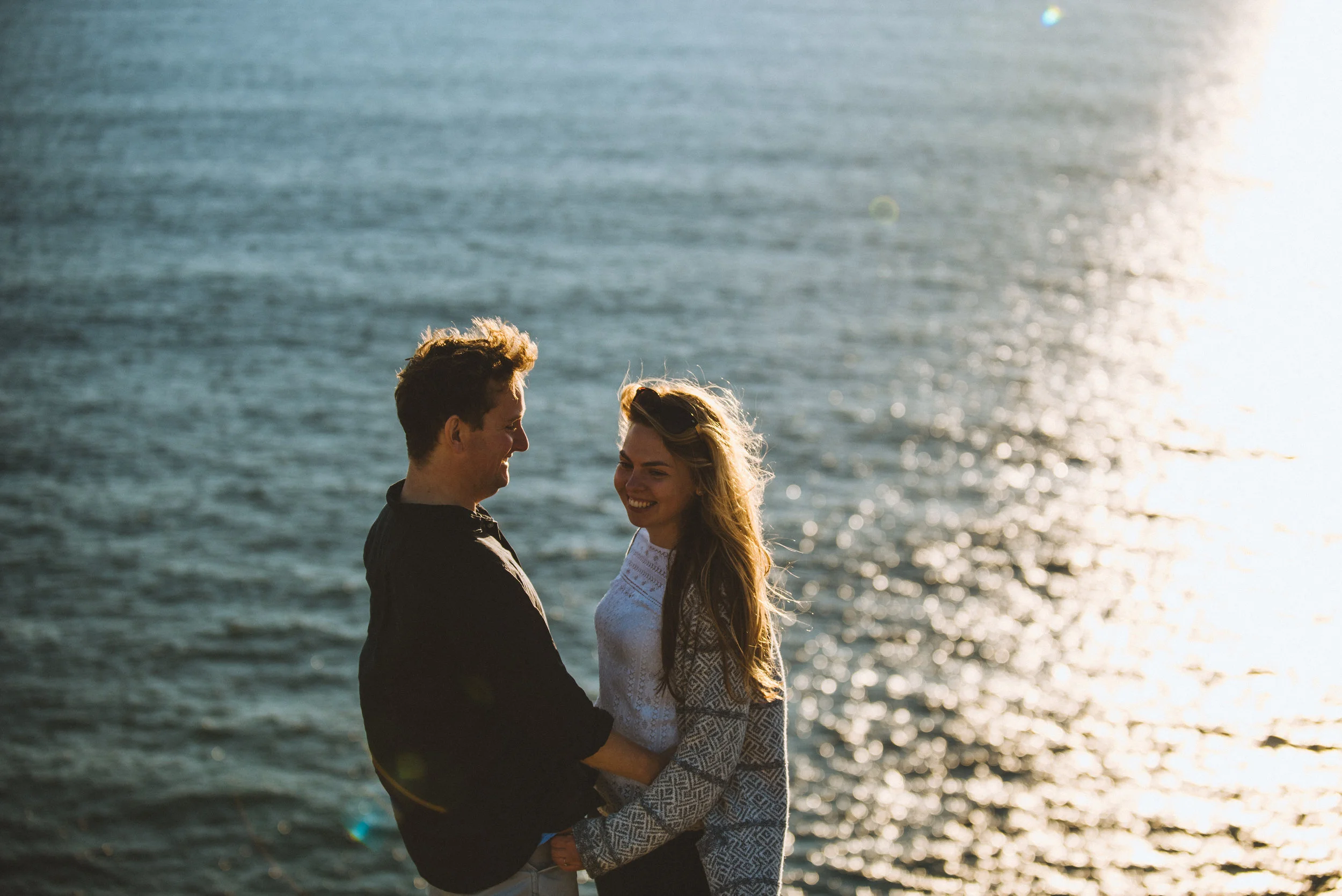 A young couple standing by the water, smiling and holding hands during sunset.