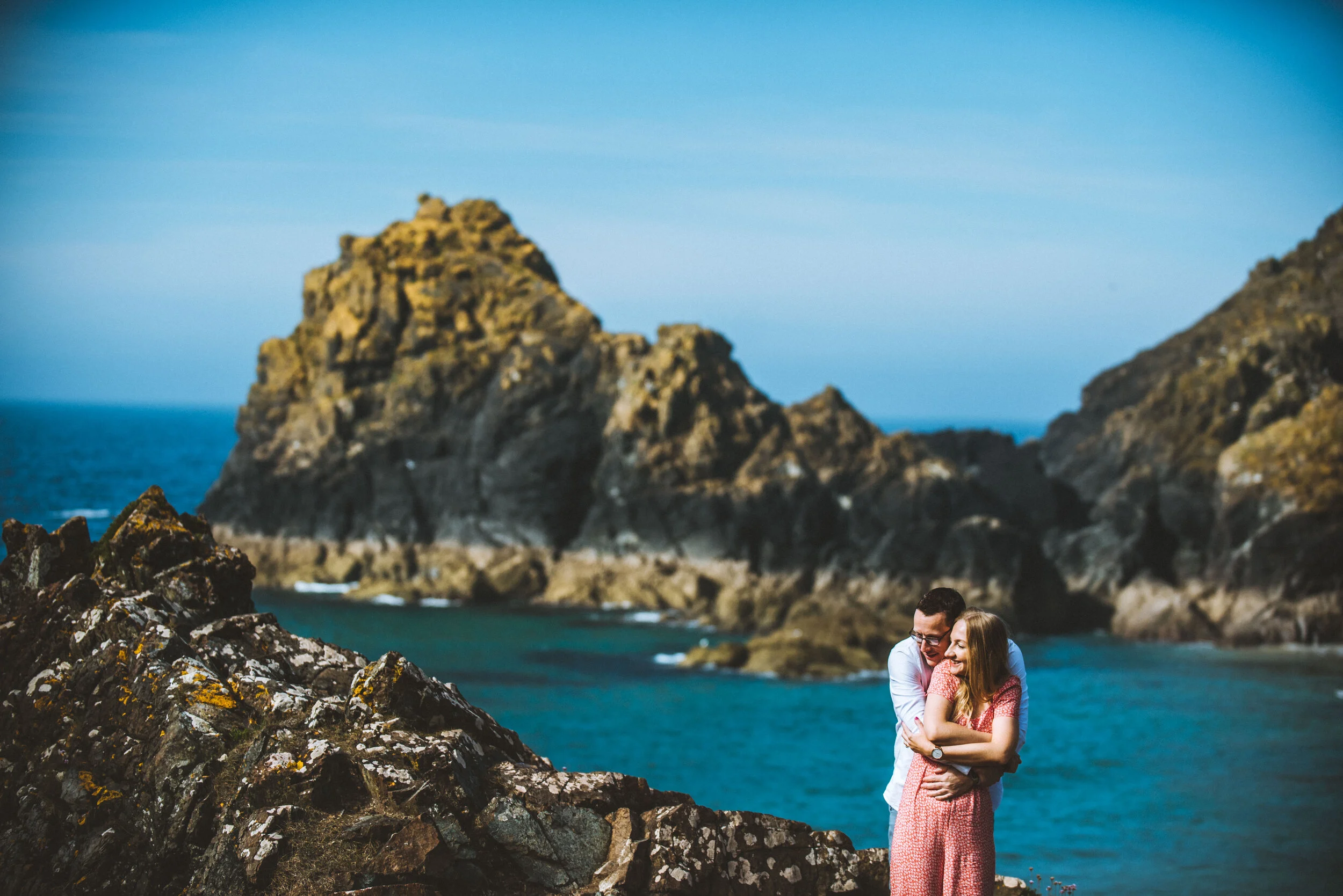 A couple hugging on a rocky shoreline with ocean and large rocky island in the background.