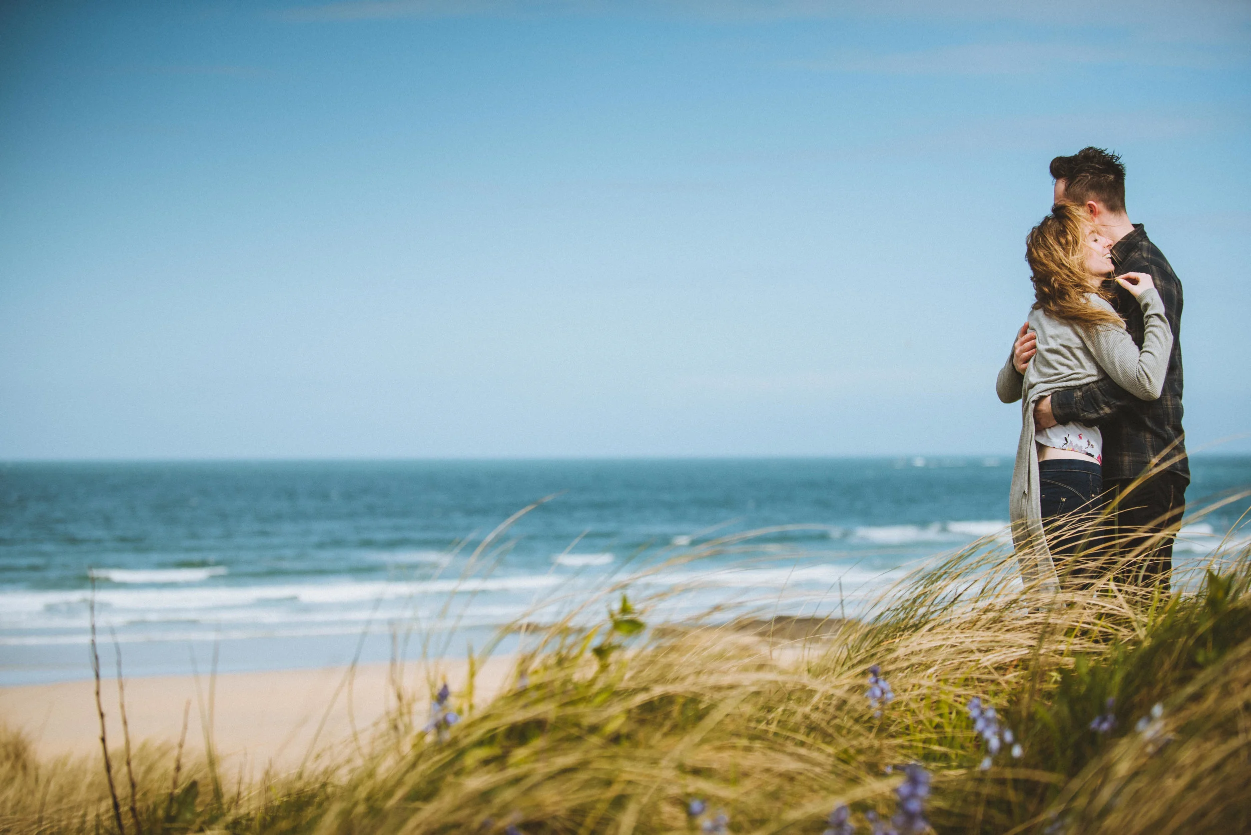 A couple hugging on a beach with grassy dunes and a blue ocean in the background.