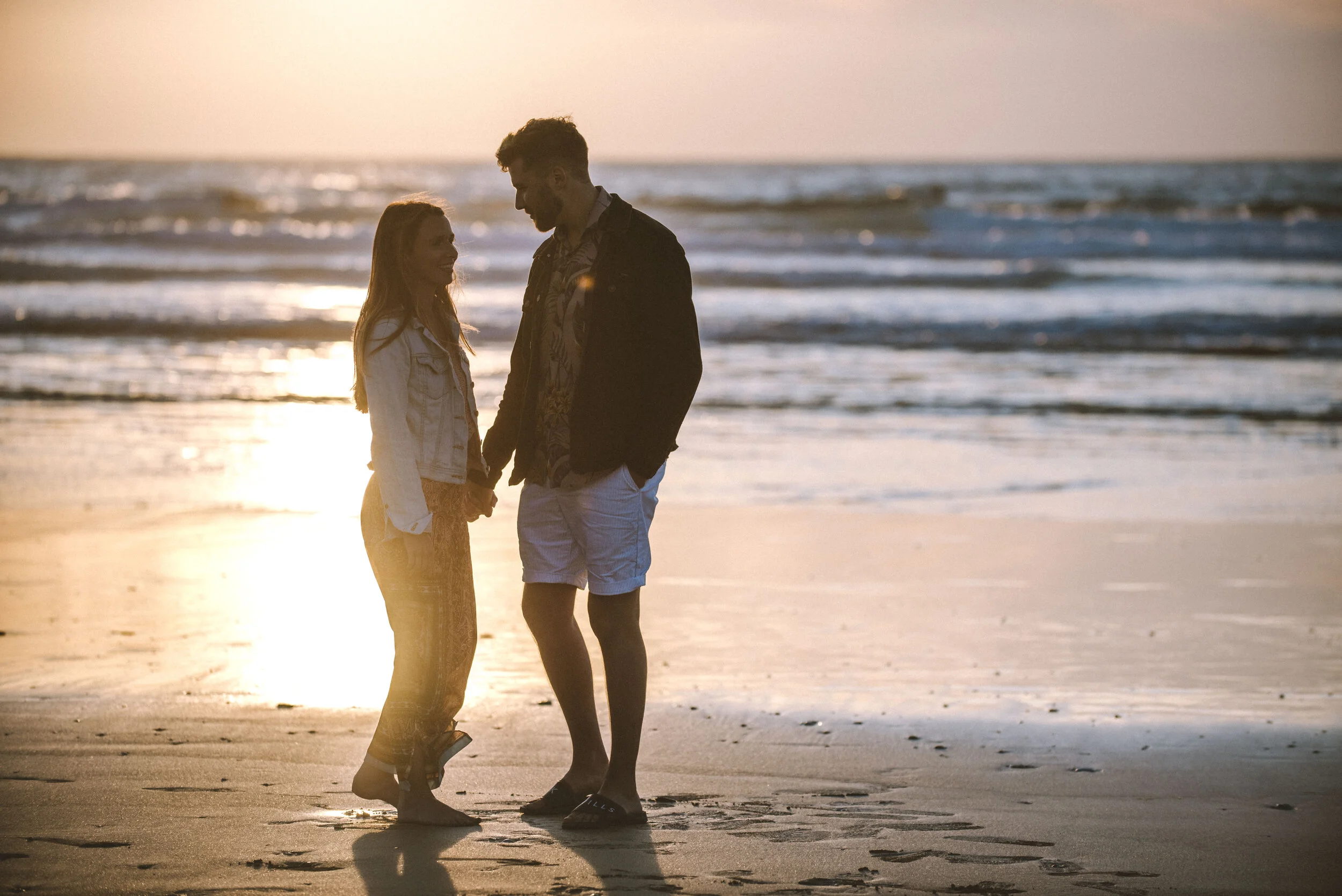 A couple holding hands on the beach at sunset, facing each other and smiling.