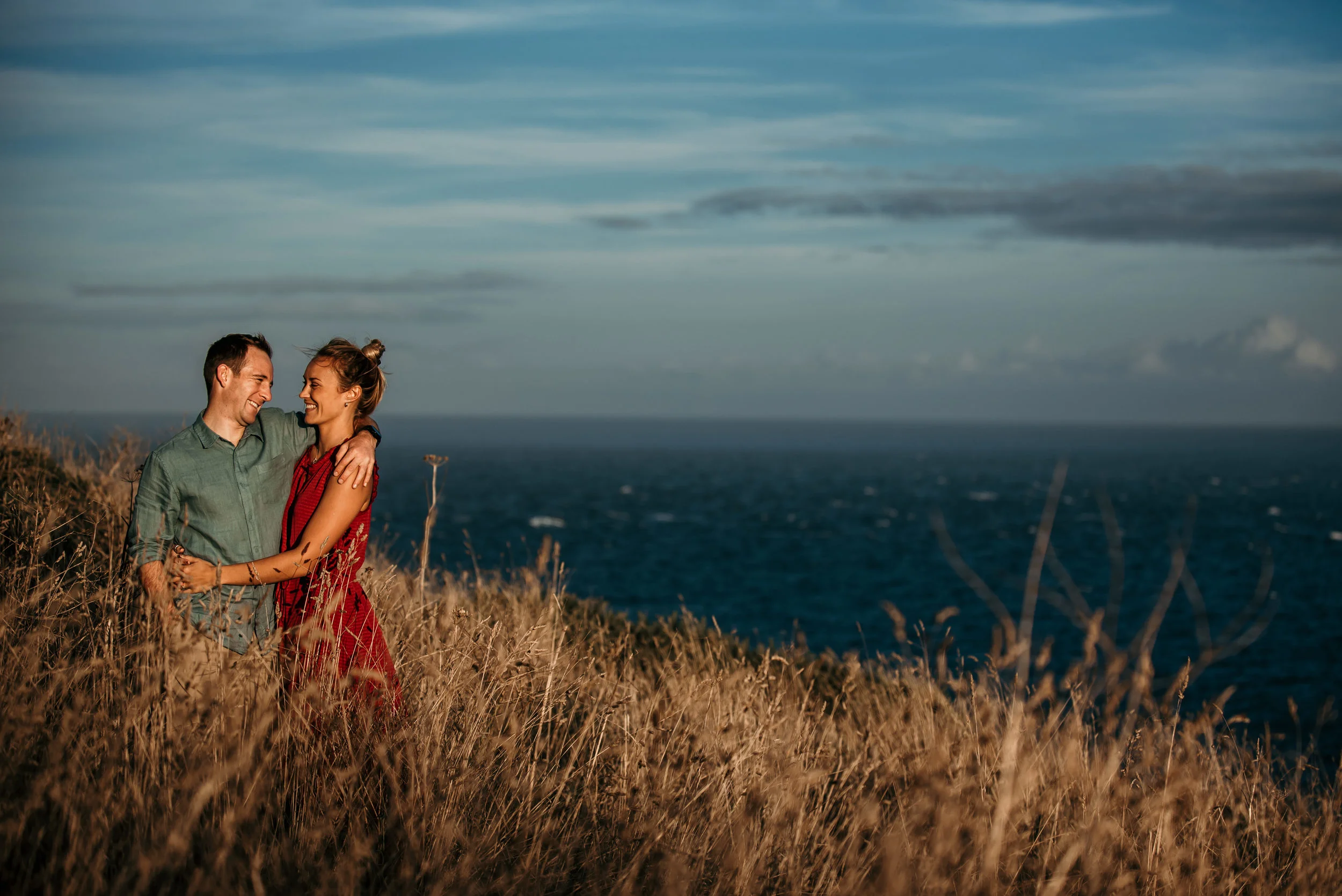 A couple standing in tall grass on a hill near the coast, smiling and embracing each other with the ocean and sky in the background.