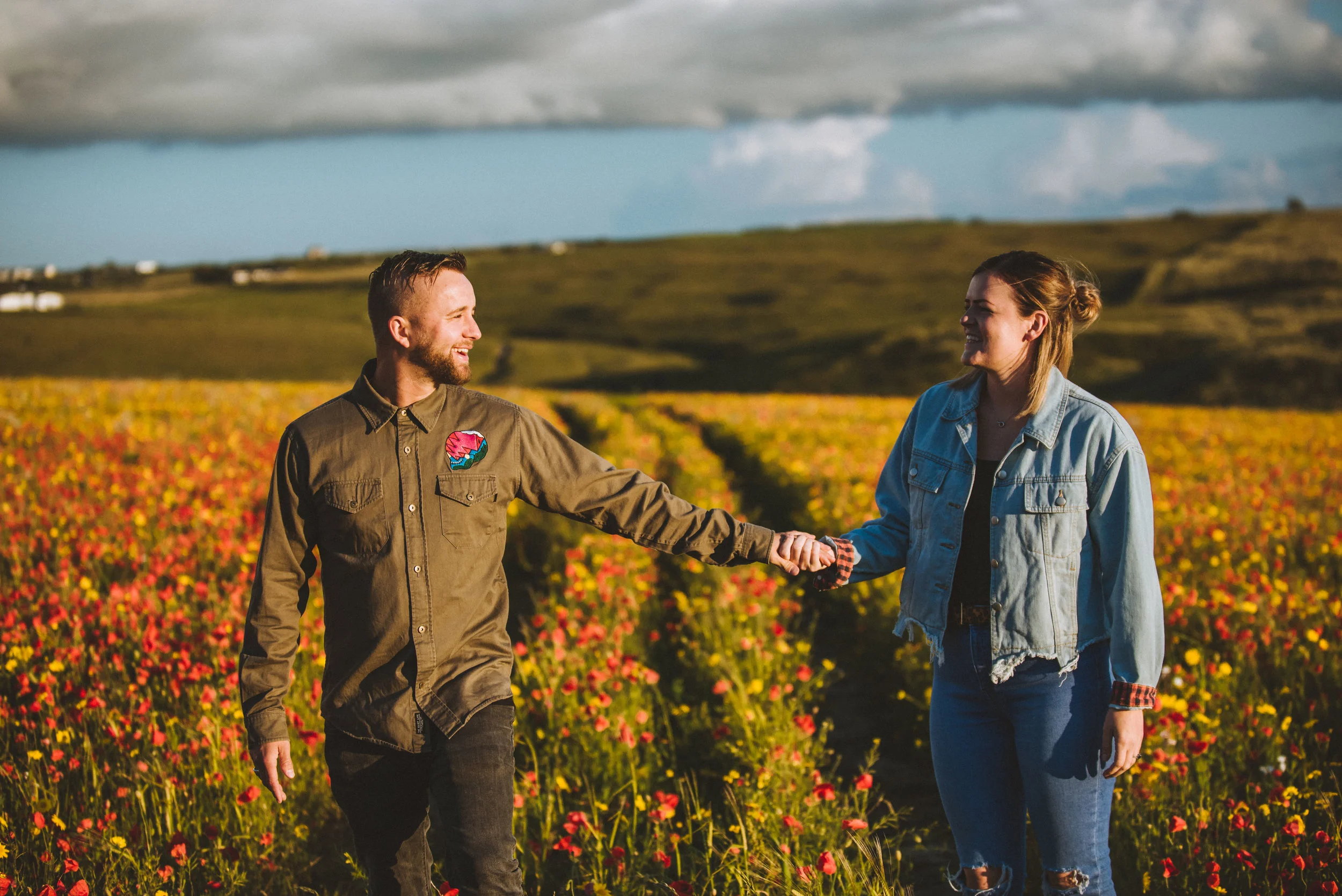 A man and woman holding hands and smiling in a field of colorful flowers with hills in the background under a partly cloudy sky.