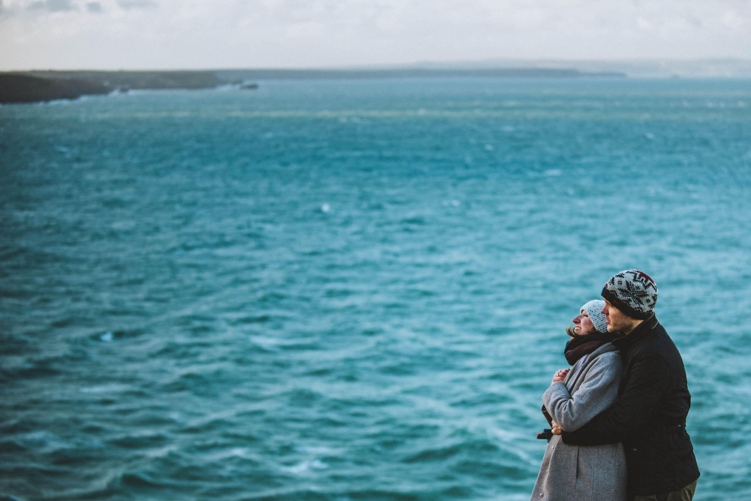 A couple standing by the water, embracing and looking out at the ocean with a distant shoreline in the background.