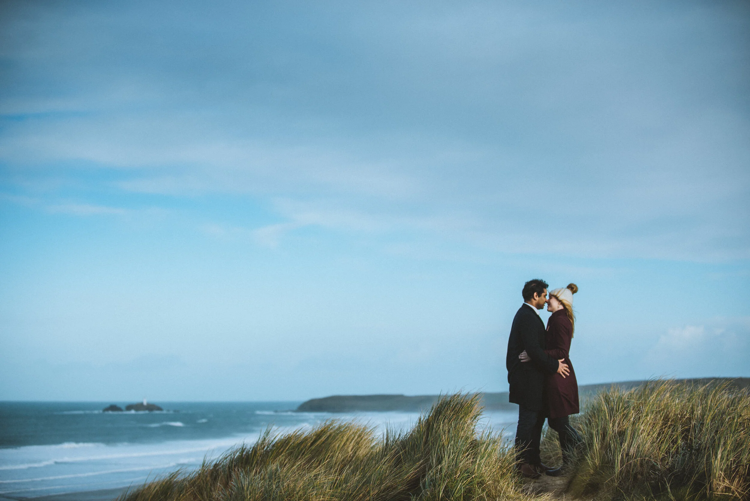 A couple standing close together on a grassy hill by the ocean, facing each other and smiling, with a blue sky and distant land in the background.