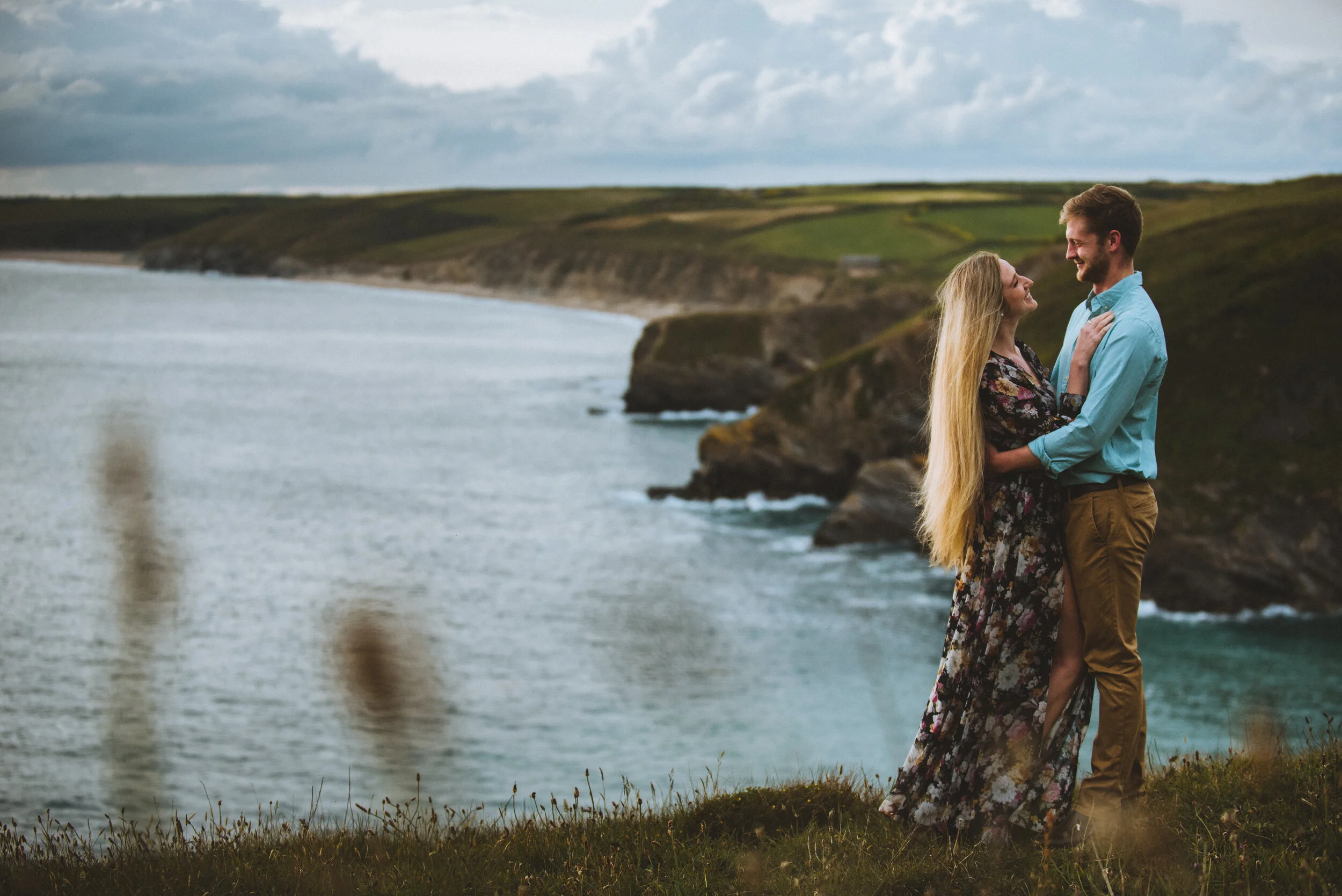 A couple standing on a grassy cliff near the ocean, embracing and smiling at each other with a cloudy sky and coastline in the background.