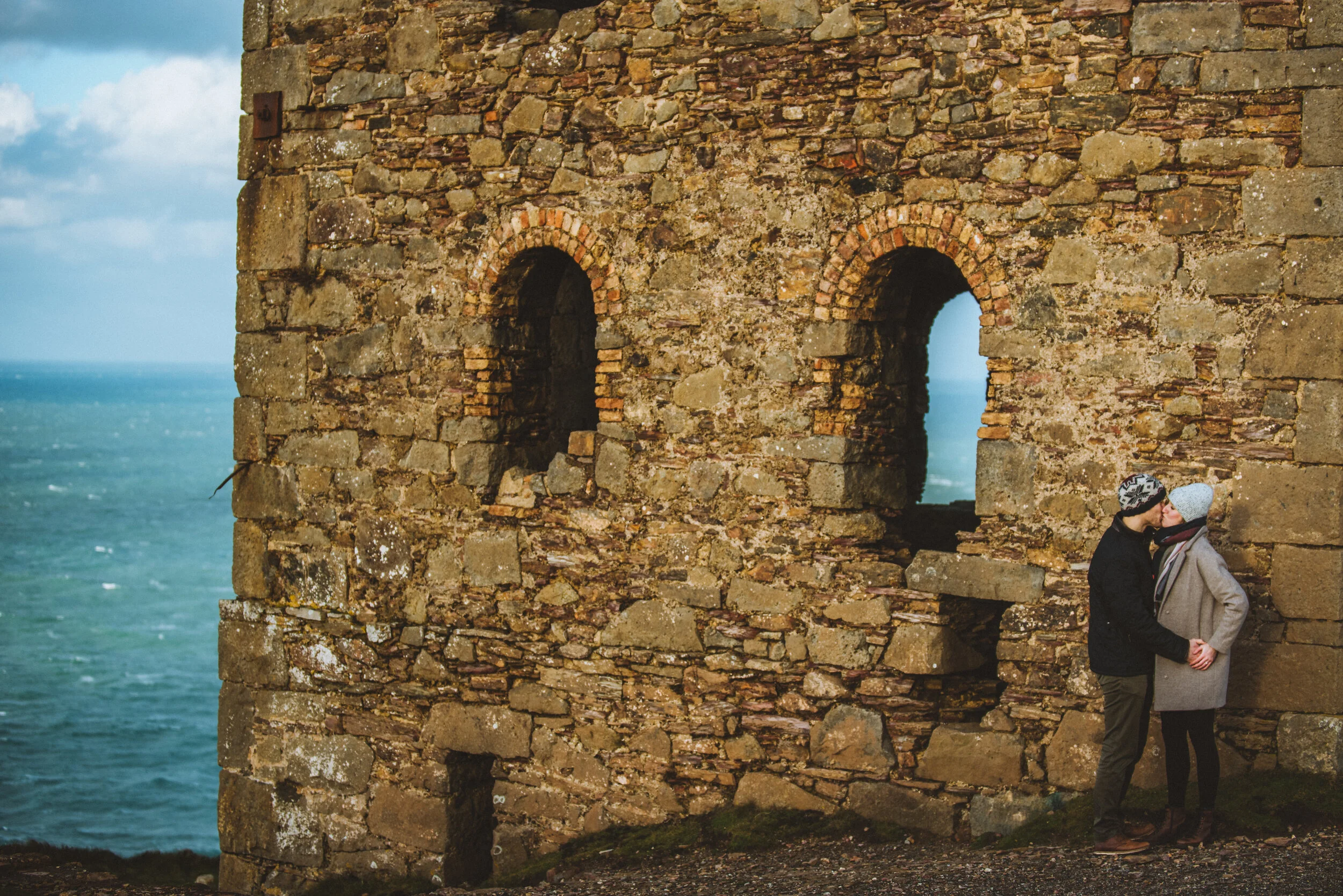Two people sharing a kiss and holding hands near an old stone building with arched windows overlooking the ocean.