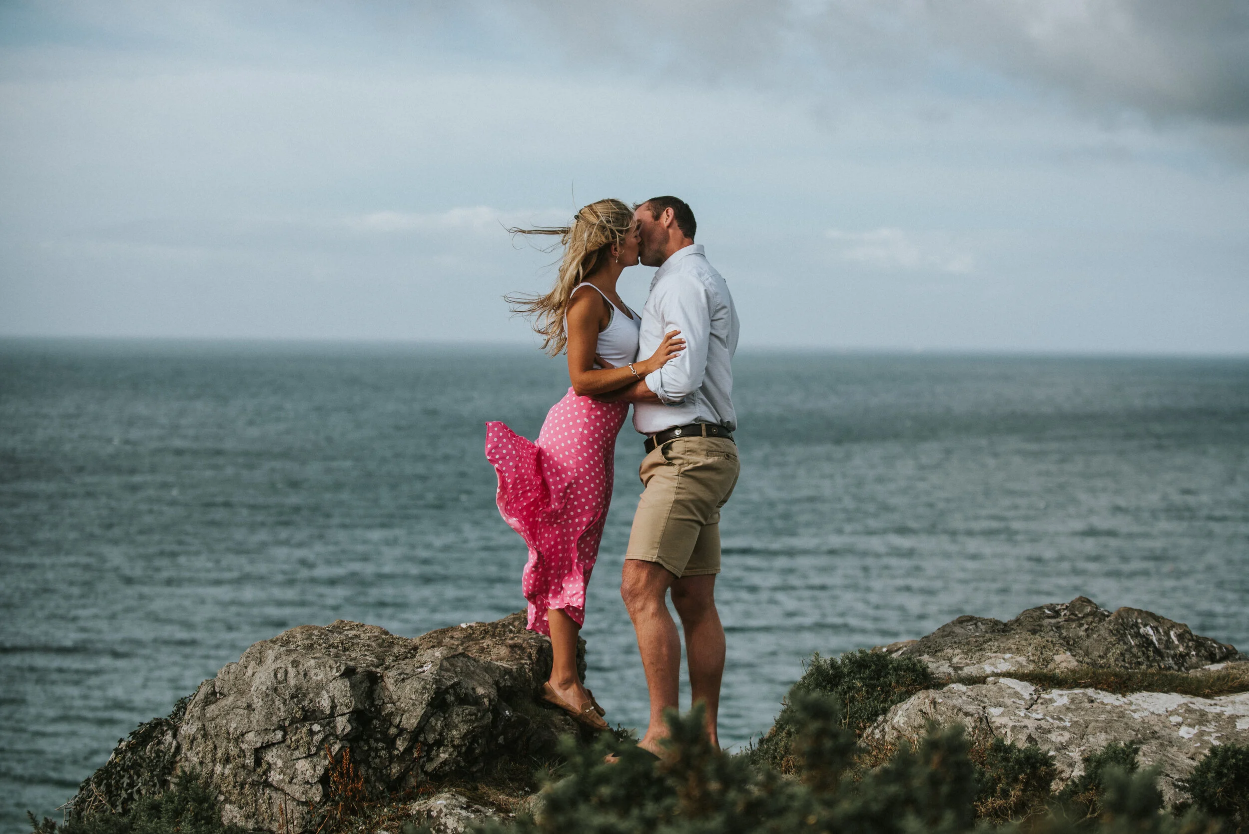 A couple is kissing on a rocky cliff overlooking the ocean during cloudy weather, with the woman wearing a pink polka dot skirt and the man in a white shirt and khaki shorts.
