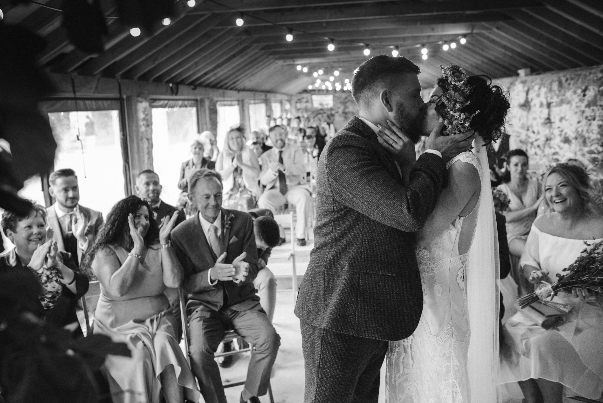 Pengenna manor wedding photography captured by Cornwall wedding photographer Mark Shaw Photography - A black-and-white photo of a wedding ceremony with a couple sharing a kiss as guests watch and applaud.