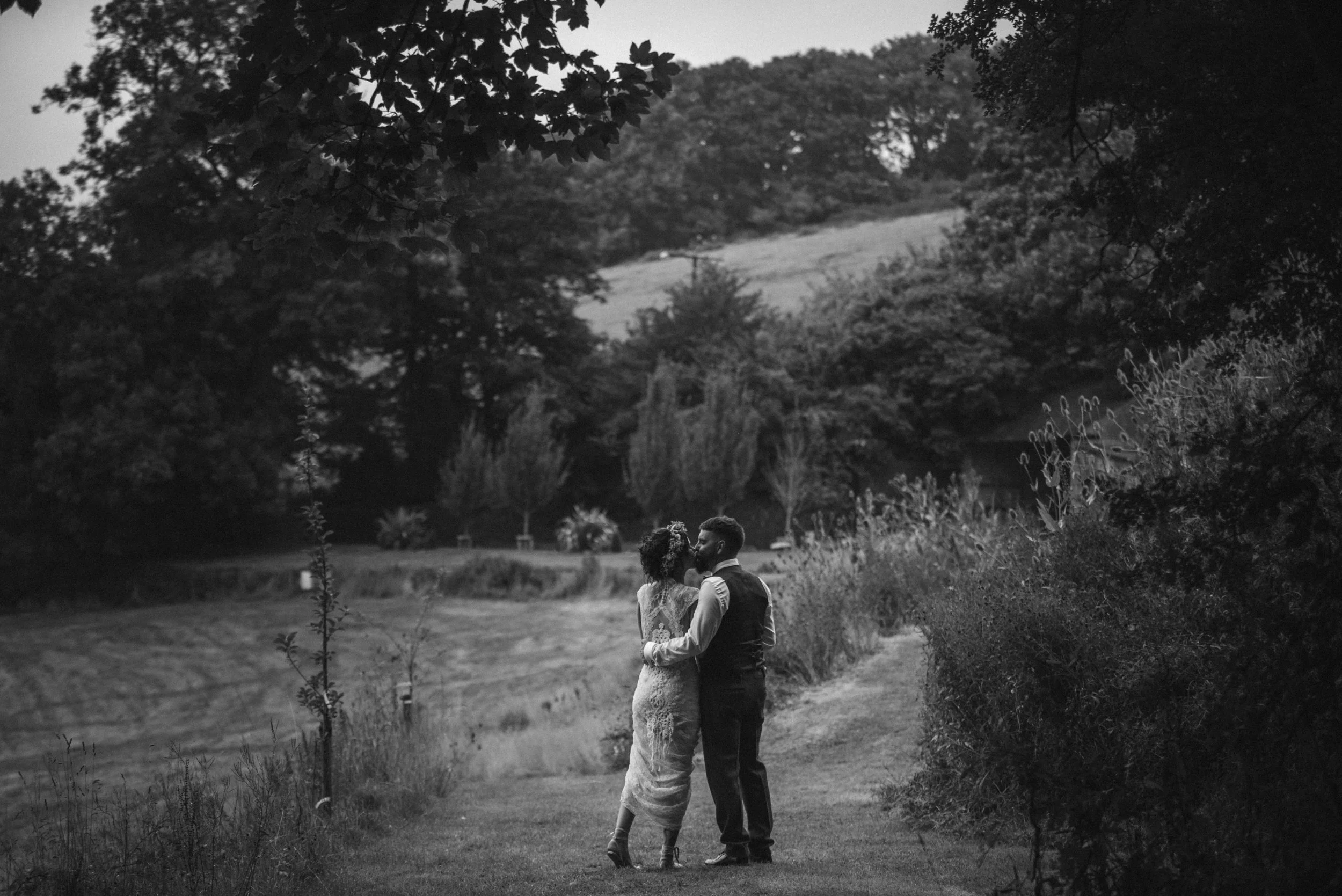 Pengenna manor wedding photography captured by Cornwall wedding photographer Mark Shaw Photography - A black-and-white photo of a couple standing closely together on a grassy path in a rural outdoor setting, surrounded by trees and bushes.