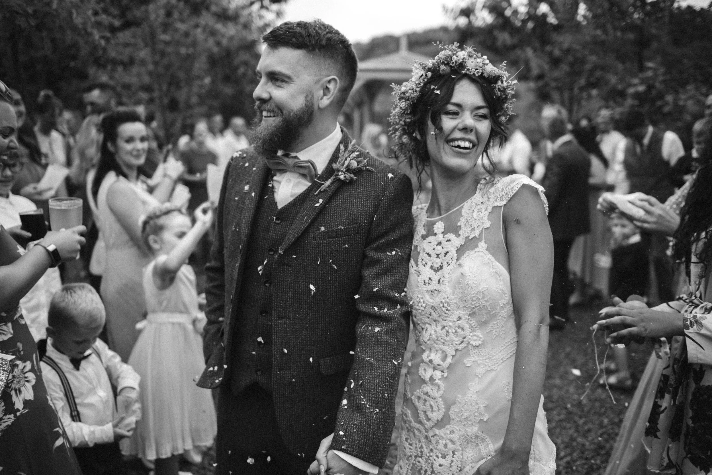 Pengenna manor wedding photography captured by Cornwall wedding photographer Mark Shaw Photography - A black-and-white photo of a wedding celebration featuring a smiling bride and groom holding hands, surrounded by guests outdoors.