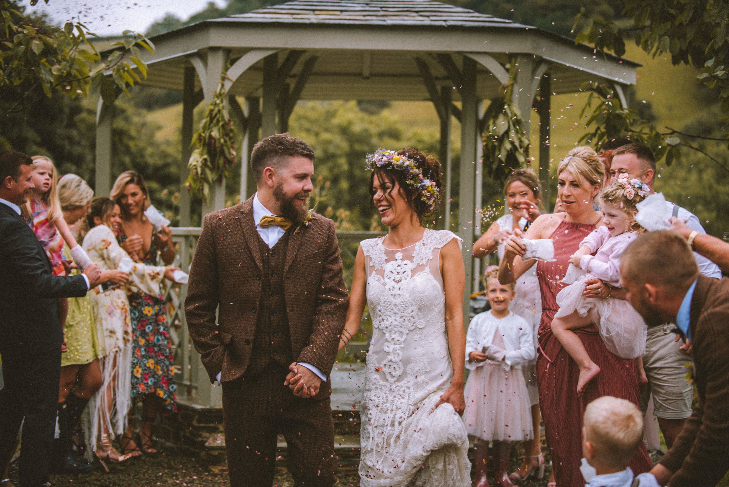 A bride and groom holding hands and walking out of a wedding ceremony outdoors, surrounded by family and friends, with confetti falling around them.