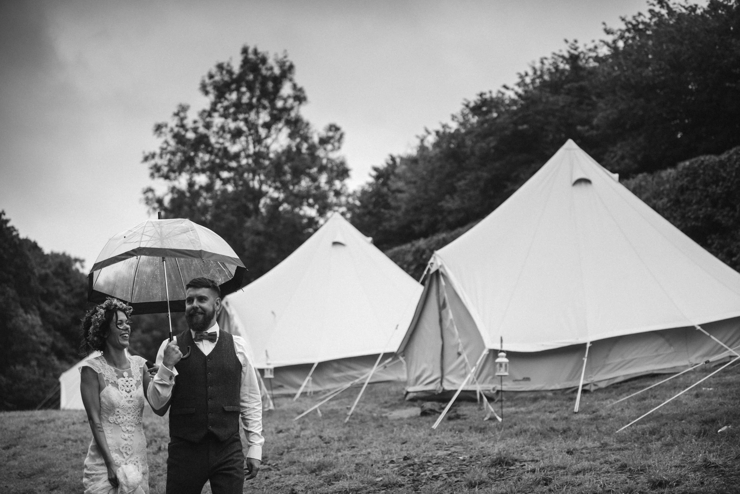 Pengenna manor wedding photography captured by Cornwall wedding photographer Mark Shaw Photography - A couple, dressed in wedding attire, walking outdoors under an umbrella near large tents on a cloudy day.
