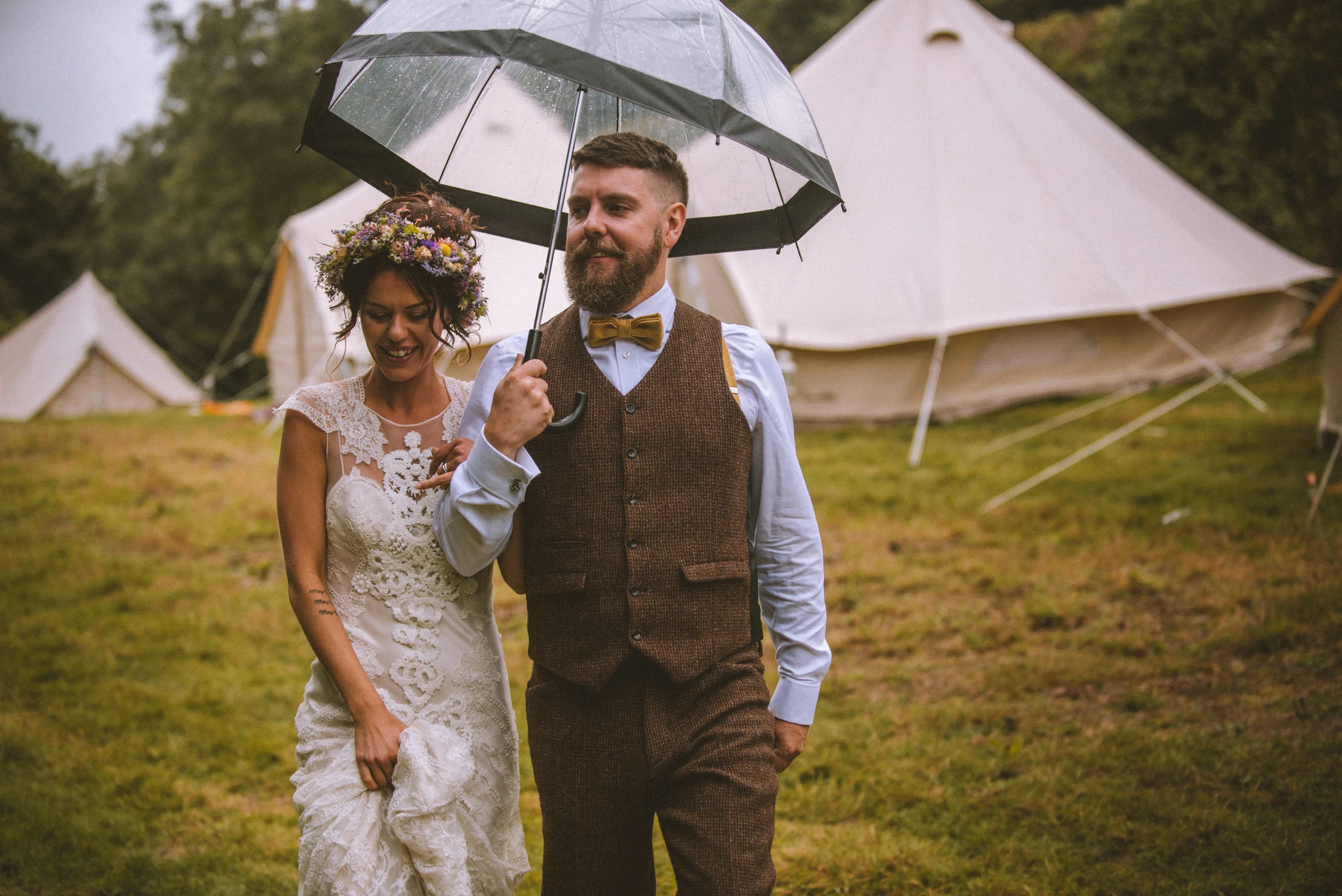 Pengenna manor wedding photography captured by Cornwall wedding photographer Mark Shaw Photography - A couple walking outside under a black umbrella, with tents in the background, during a rainy outdoor event.