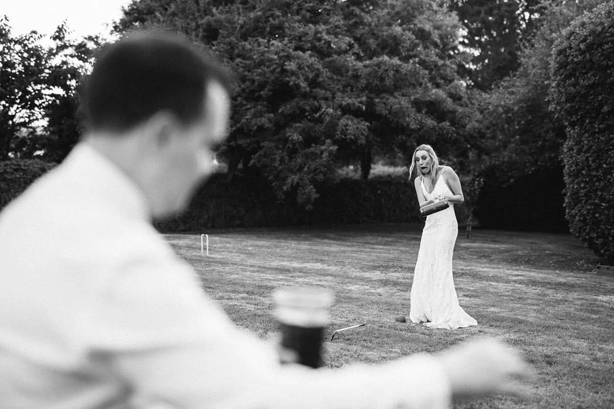 A woman in a white dress holding a tray stands on a lawn, looking surprised, while a man in glasses is out of focus in the foreground near a cup.