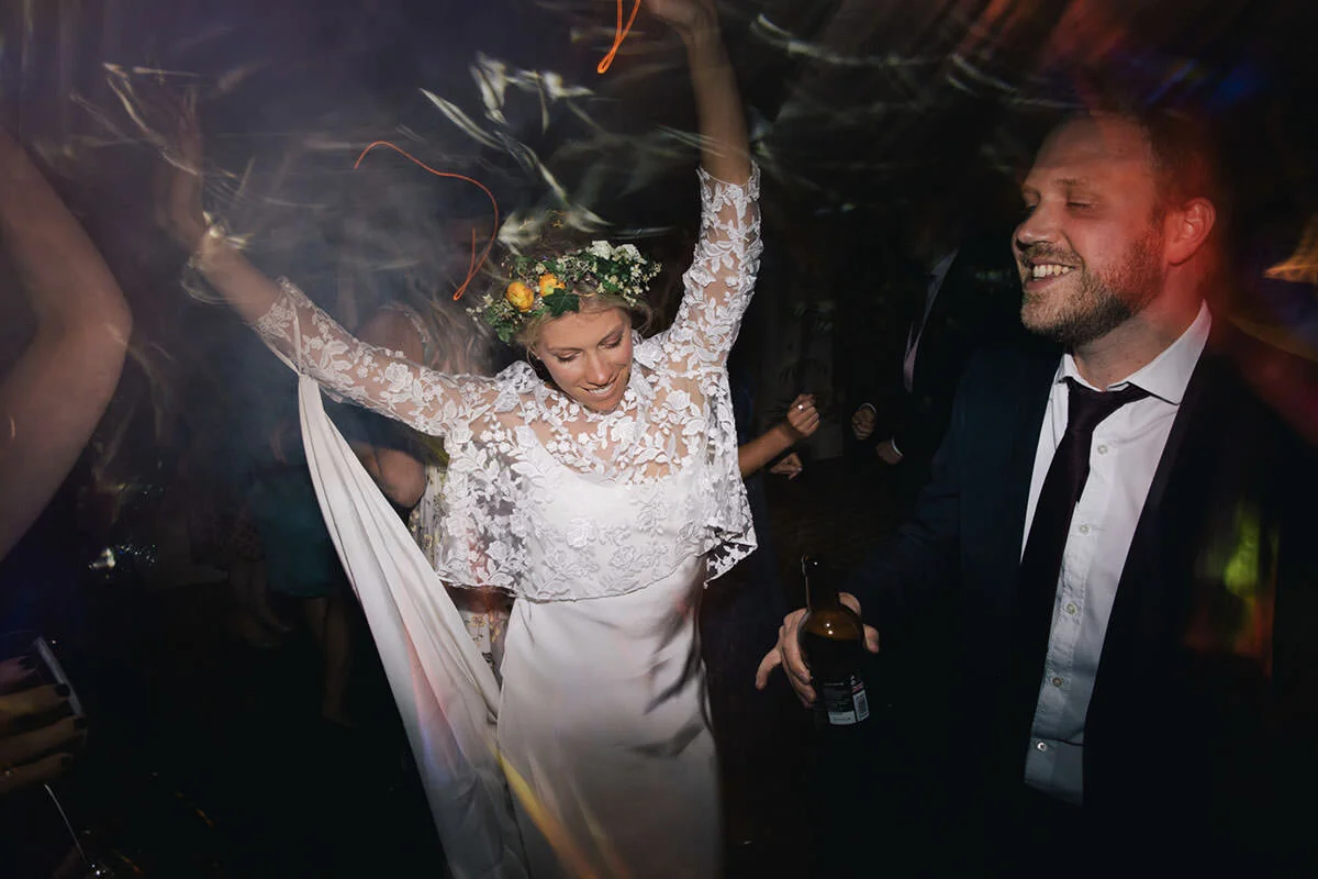 A bride wearing a floral crown and lace wedding dress dancing and smiling at her wedding reception, while a man in a suit holds a beer and dances nearby.