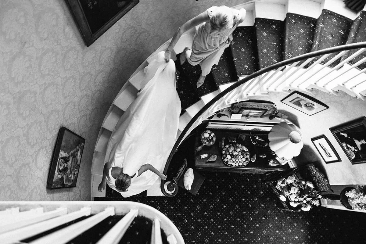 A woman in a wedding dress walks up a curved staircase inside a house, seen from above in black and white.