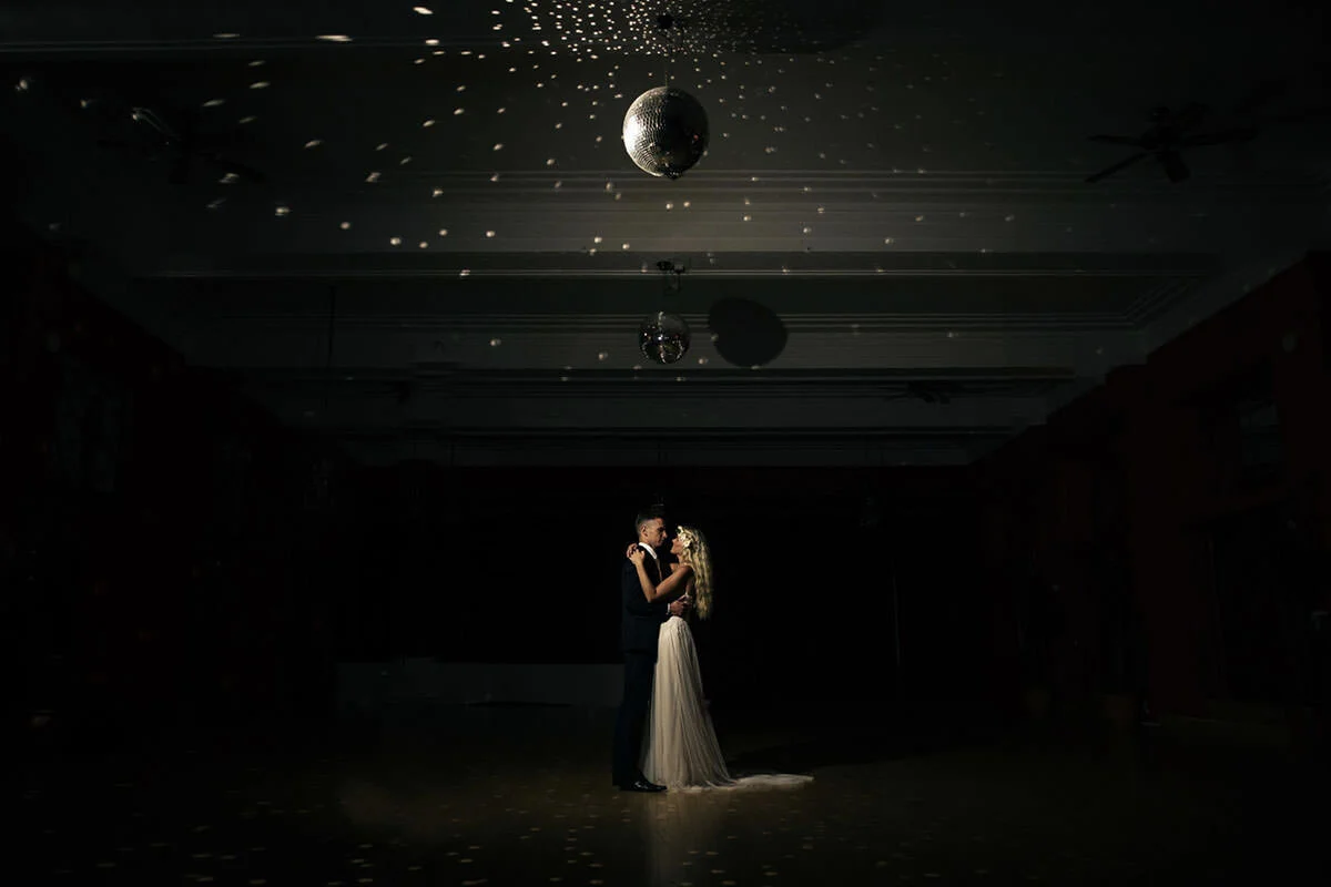 A couple in wedding attire dancing under a disco ball in a dimly lit room.
