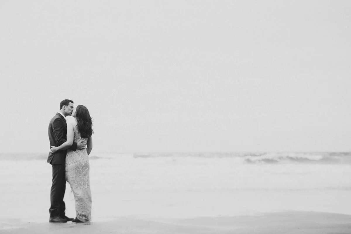 A black and white photo of a man and woman on the beach, standing close together and kissing.