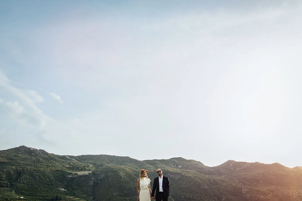 A couple dressed in wedding attire walking hand in hand in a scenic outdoor landscape with green hills and a partly cloudy sky.