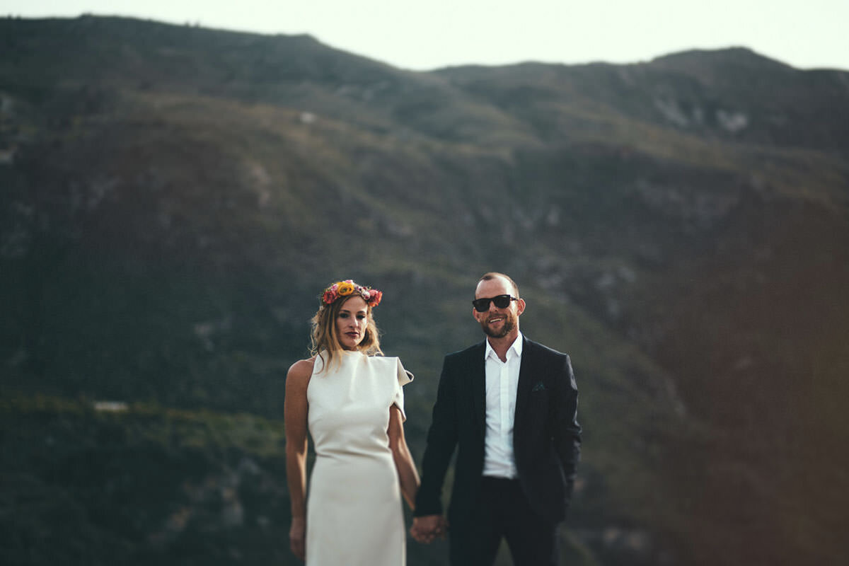 A man and woman in wedding attire holding hands outdoors with mountains in the background.