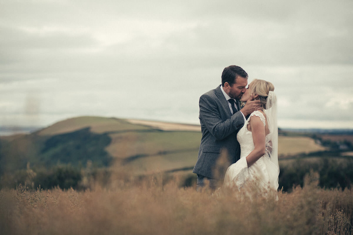 A man and woman dressed in wedding attire sharing a kiss in an outdoor rural setting with rolling hills in the background.