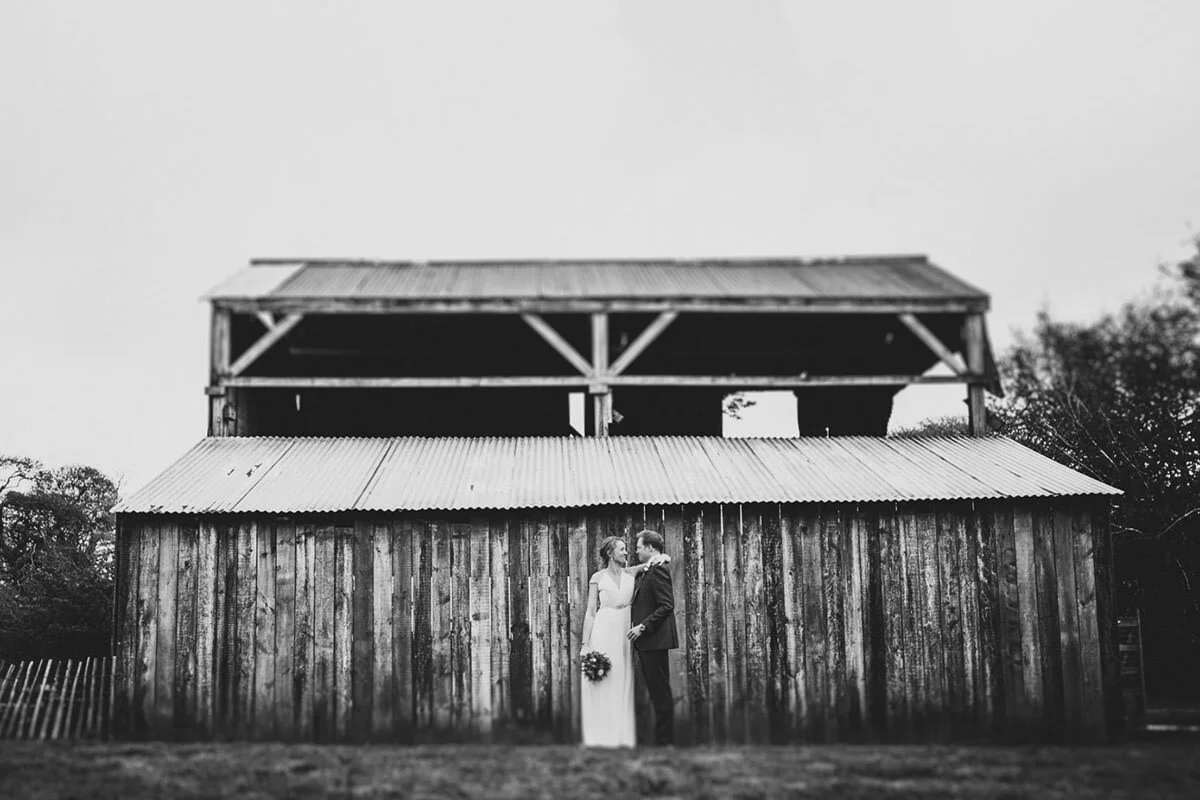 A bride and groom standing close, holding hands, and looking at each other in front of a rustic wooden barn.