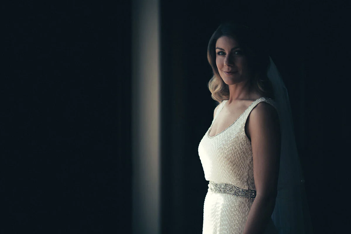 A woman in an elegant white wedding dress with a silver belt, standing in a dimly lit room and looking at the camera with a slight smile.