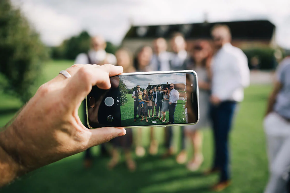 A hand holding a smartphone taking a photo of a group of people outdoors on a green lawn with a large house in the background.