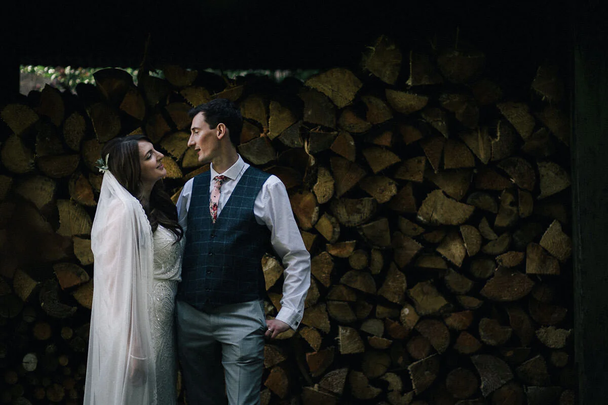 A couple dressed in wedding attire looking at each other in front of a large stack of cut firewood.