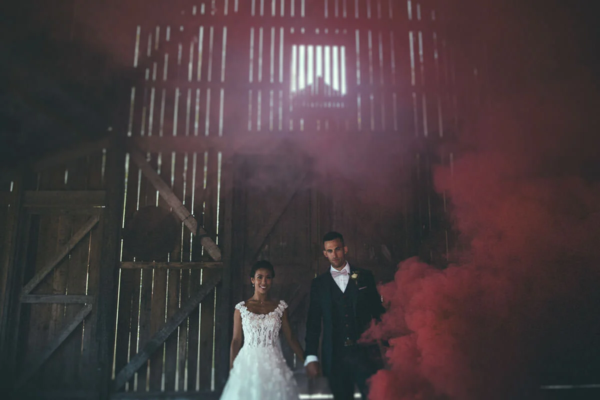 A bride and groom walking hand in hand inside a barn with pink smoke and sunlight filtering through wooden slats.