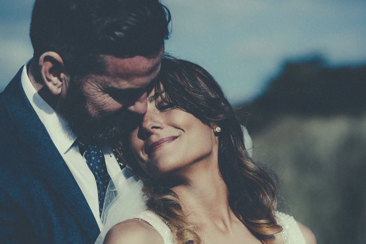 A man in a suit and a woman in a white dress sharing a tender moment outdoors, with the man kissing the woman's forehead as she smiles.