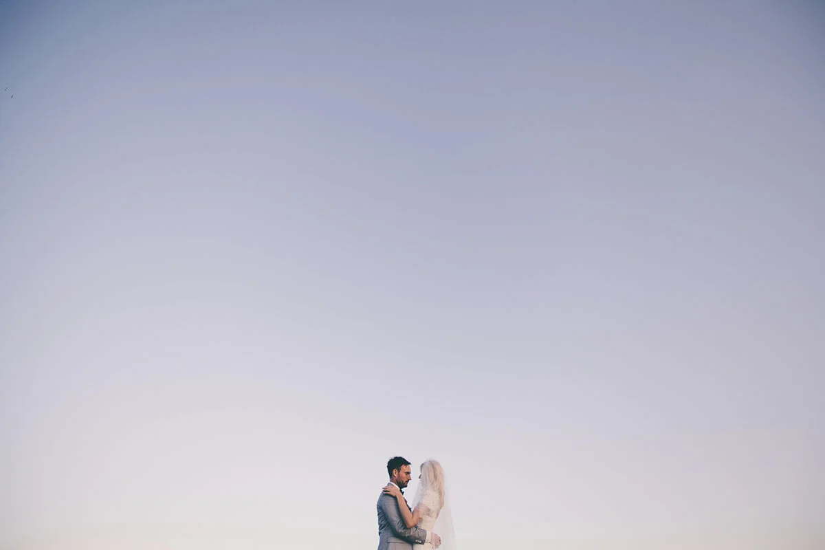 A couple, dressed in wedding attire, faces each other against a vast, clear sky.
