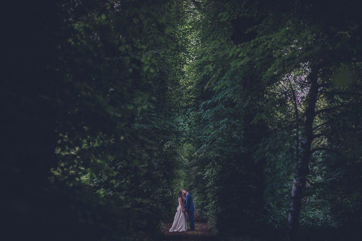 A bride and groom standing close together in a lush, green forest with tall trees forming a natural canopy overhead.