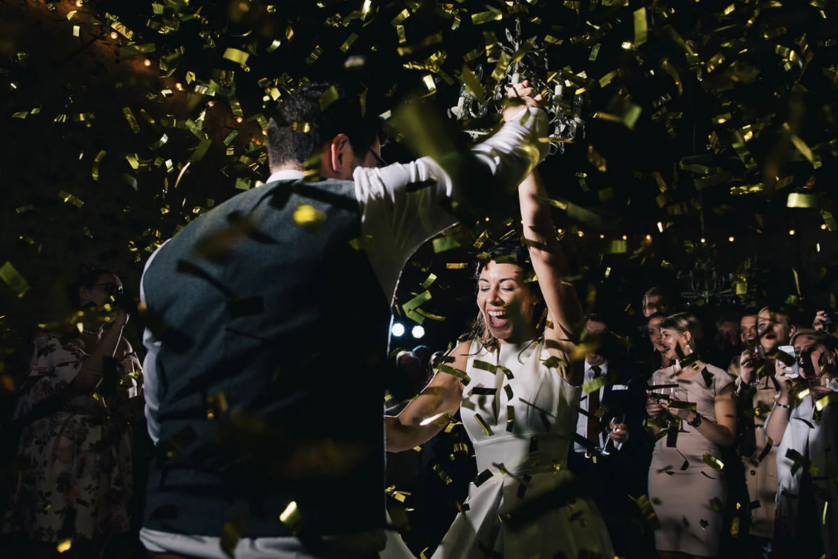 People celebrating with gold confetti, a woman dancing with a joyful expression, surrounded by others watching and taking photos at an indoor event.