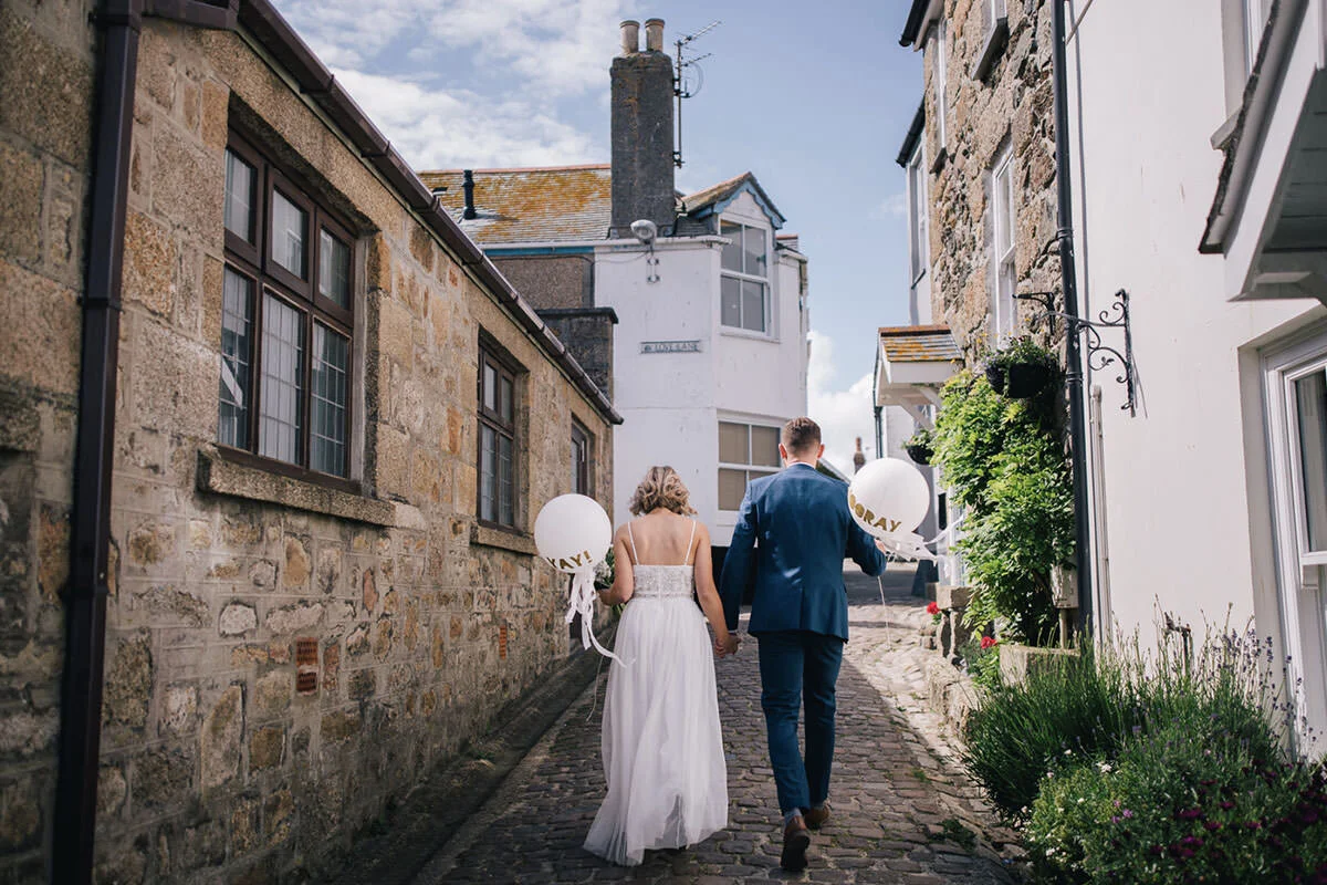 A couple dressed in wedding attire walking hand in hand down a cobblestone alleyway, carrying balloons that say "hooray" for their celebration, surrounded by old stone and white buildings with greenery.