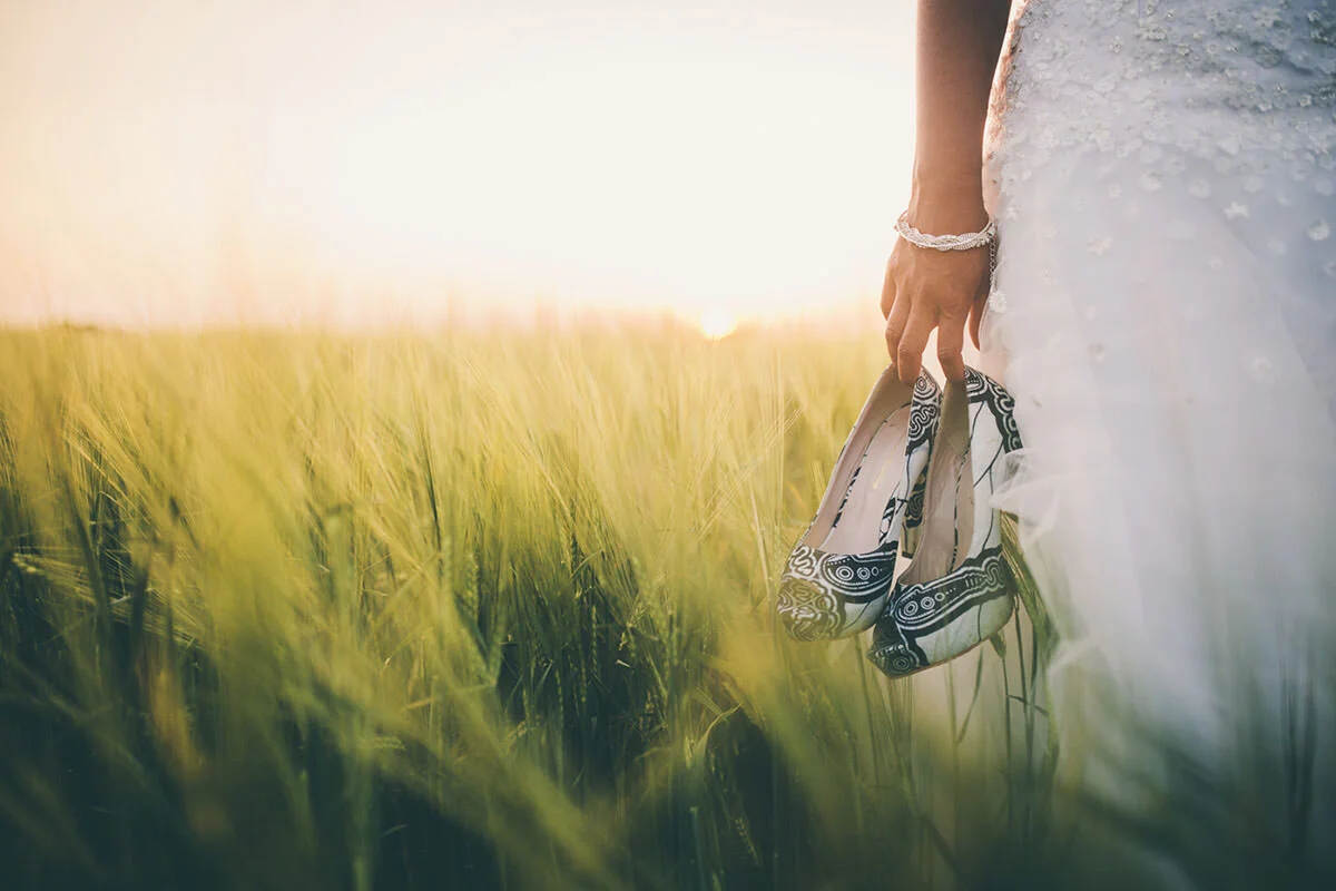 A bride standing in a field at sunset, holding a pair of patterned shoes in one hand, with her wedding dress and a bracelet visible.