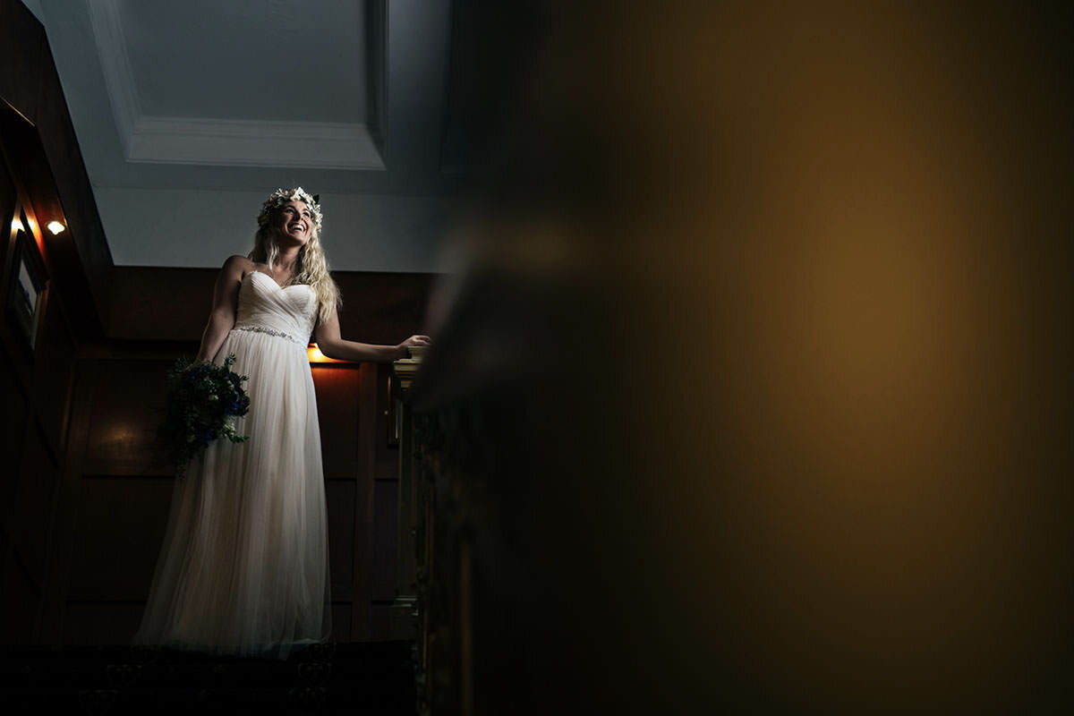 A woman wearing a wedding dress, flower crown, and holding a bouquet, smiling and standing near a stair railing in a dimly lit room.