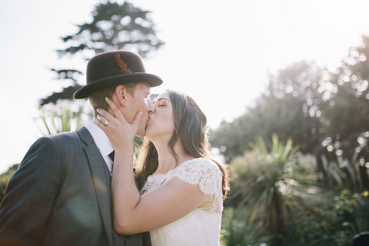A couple kissing outdoors, the man wearing a hat with a feather and a suit, the woman in a white lace dress, with greenery and trees in the background.