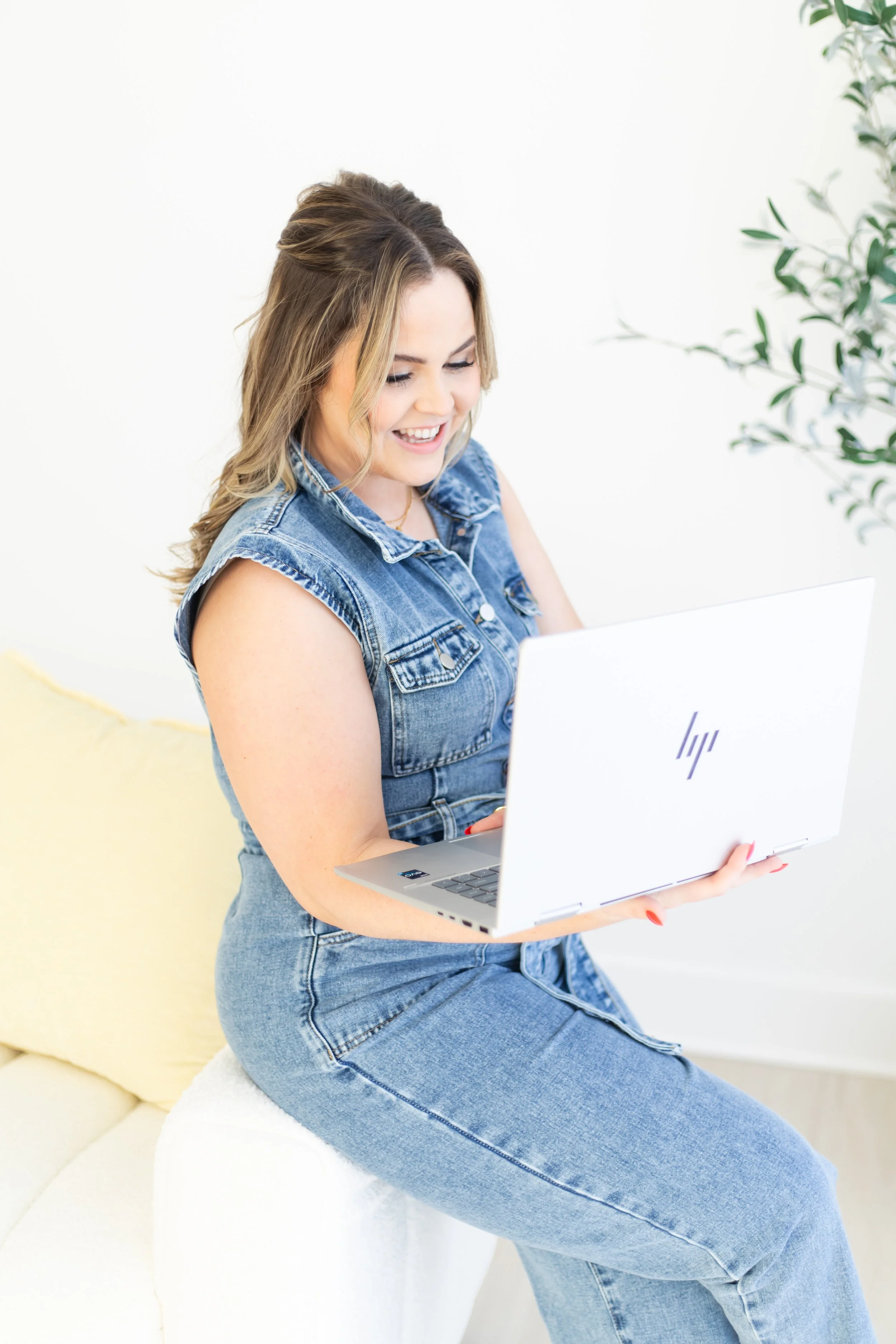 A young woman with wavy blonde hair sitting on a white cushioned seat, smiling and working on a white HP laptop, wearing a sleeveless denim vest and matching denim jeans, with a green plant in the background.