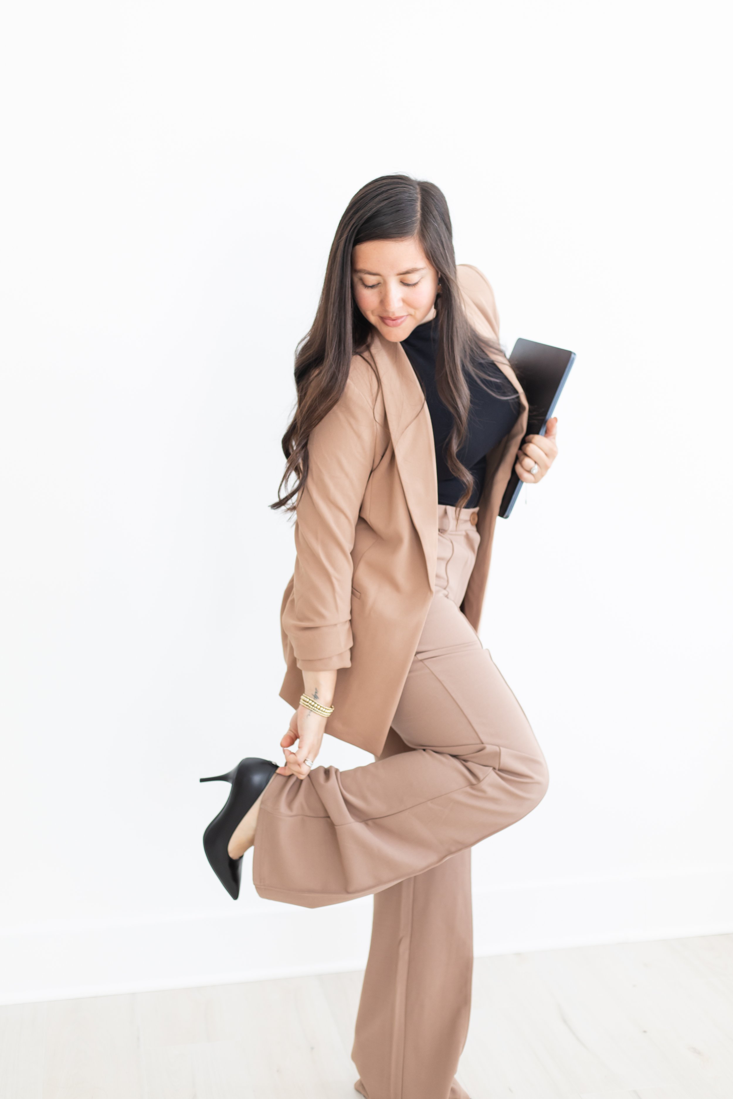 Woman in beige suit with black top and heels, holding a tablet, kneeling on one knee with her head bowed, against a plain white background.
