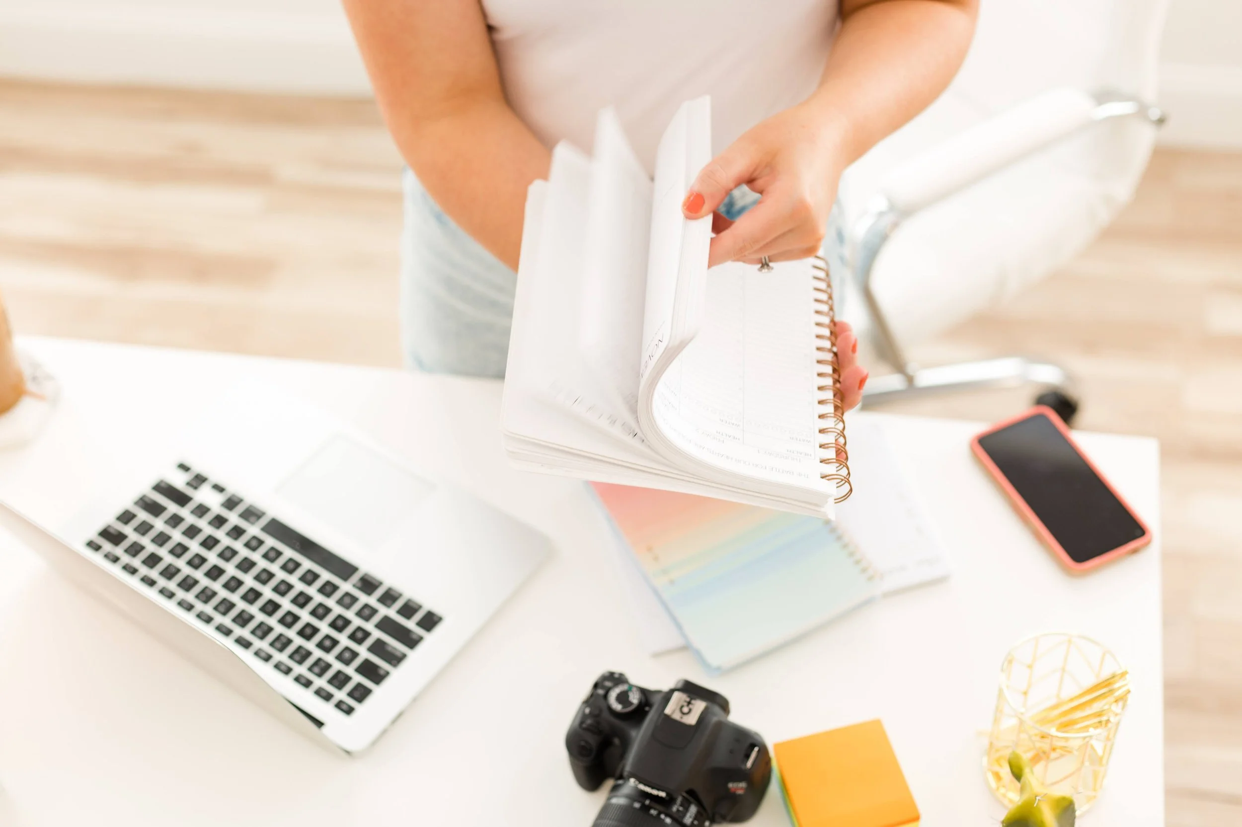 Person wearing a white top and light-colored pants holding a white spiral notebook, standing at a white desk with a laptop, a camera, a smartphone, colorful notebooks, a yellow cone with paper clips, and a glass with lemon slices and mint leaves.