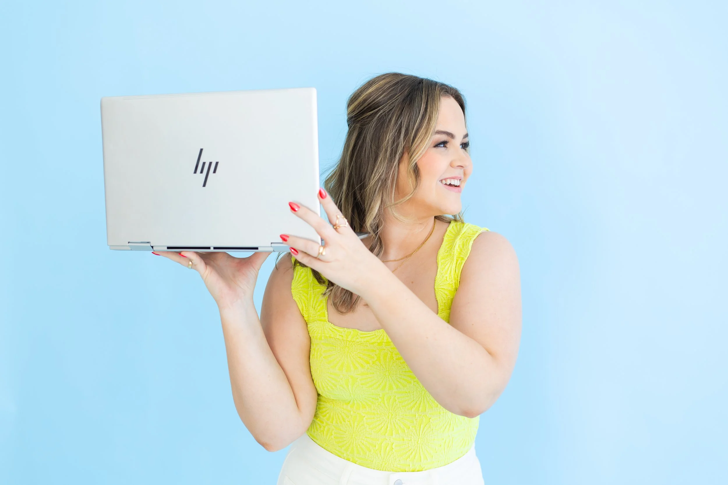 A woman with long wavy hair, wearing a bright yellow sleeveless top and white pants, smiling and looking to the side while holding a white HP laptop in front of her against a light blue background.