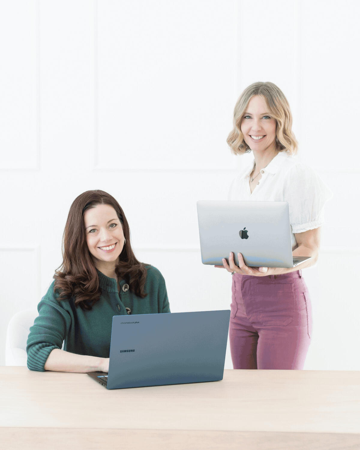 Two women working with laptops in a bright, white office space. One woman is sitting at the desk using a Samsung Chromebook, while the other is standing, holding a silver Apple MacBook.