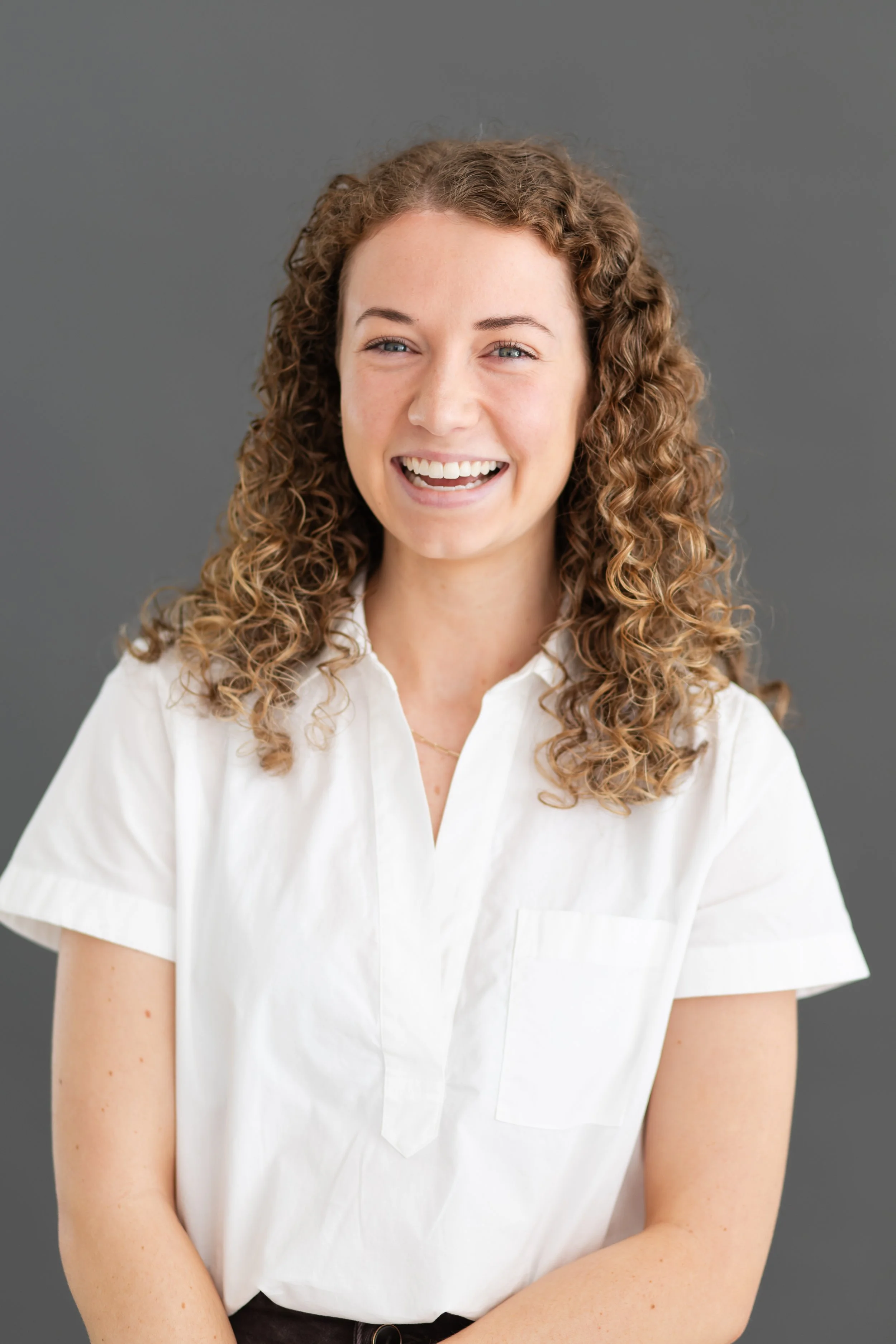 A woman with curly hair wearing a white shirt, smiling and looking at the camera against a gray background.