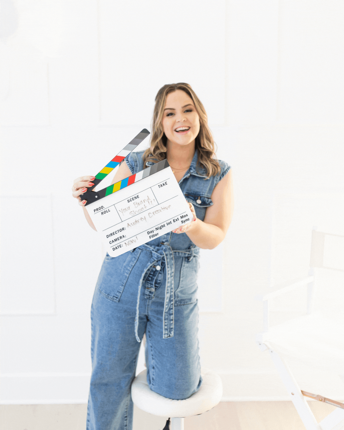 A woman in a denim outfit holding a film clapperboard, standing on a white chair in a bright room.