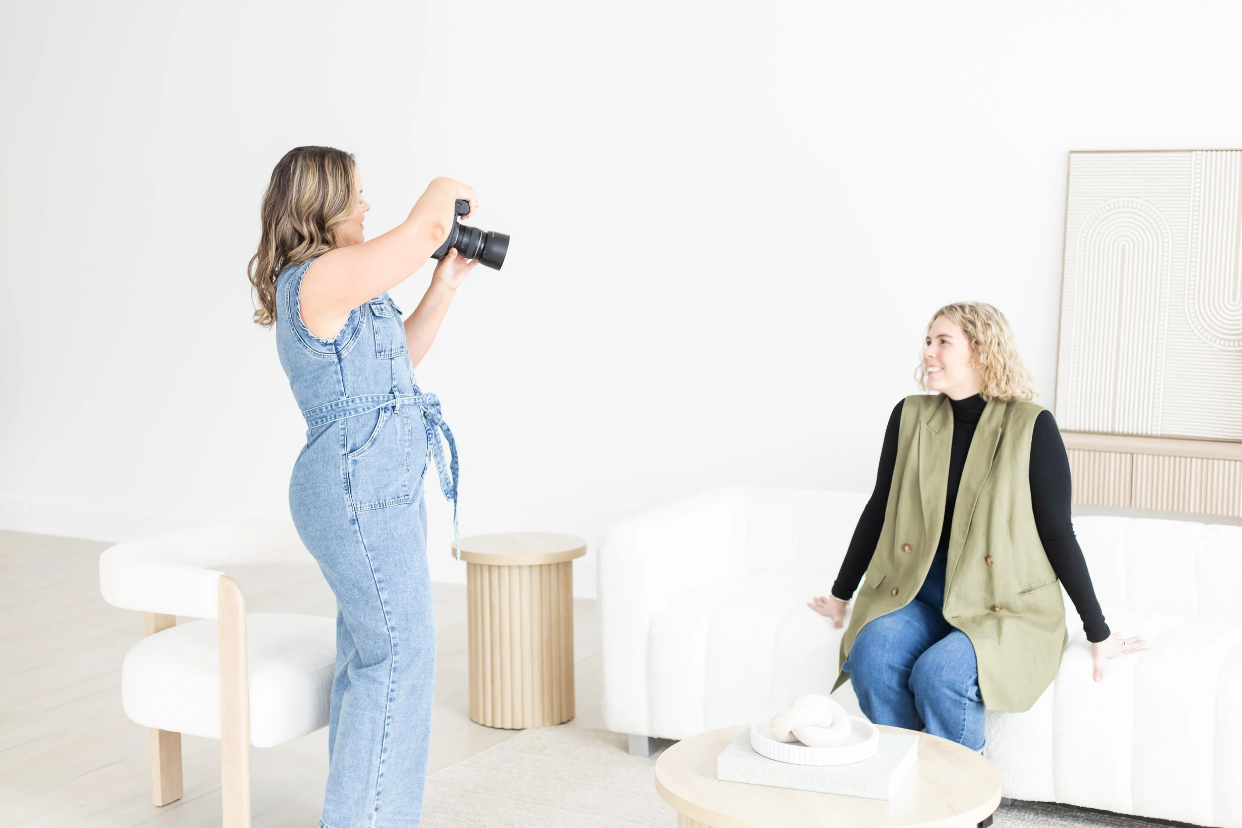 Two women inside a bright, minimalist room. One woman is standing and holding a camera, taking a photo of the other woman, who is sitting on a white chair and smiling.